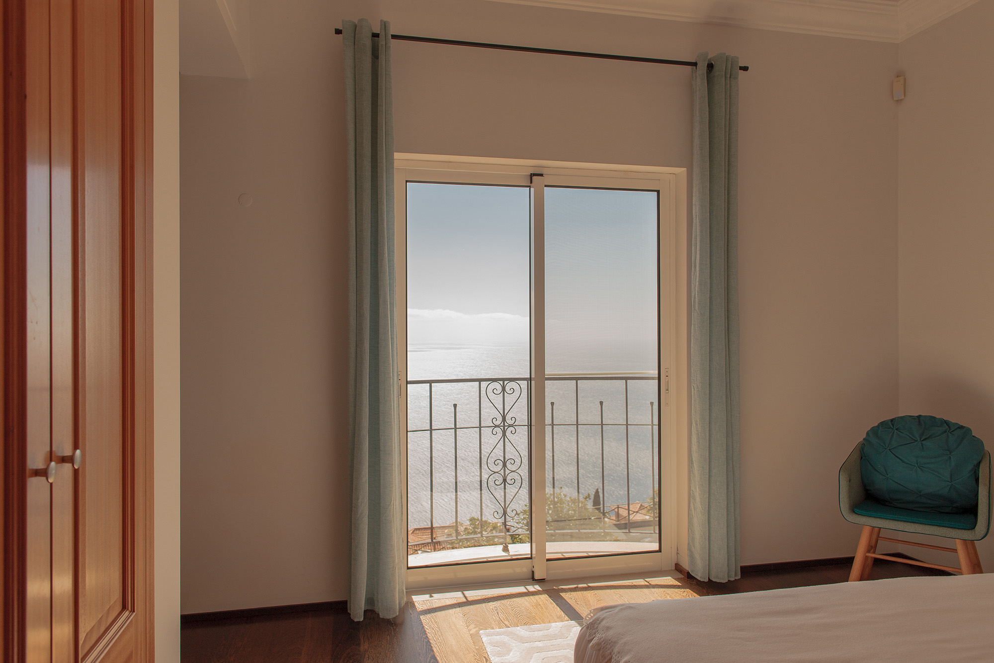 Bedroom with open sliding balcony door, teal curtains, teal armchair, wooden wardrobe, and Atlantic ocean view, Palheiro Village villa, Madeira
