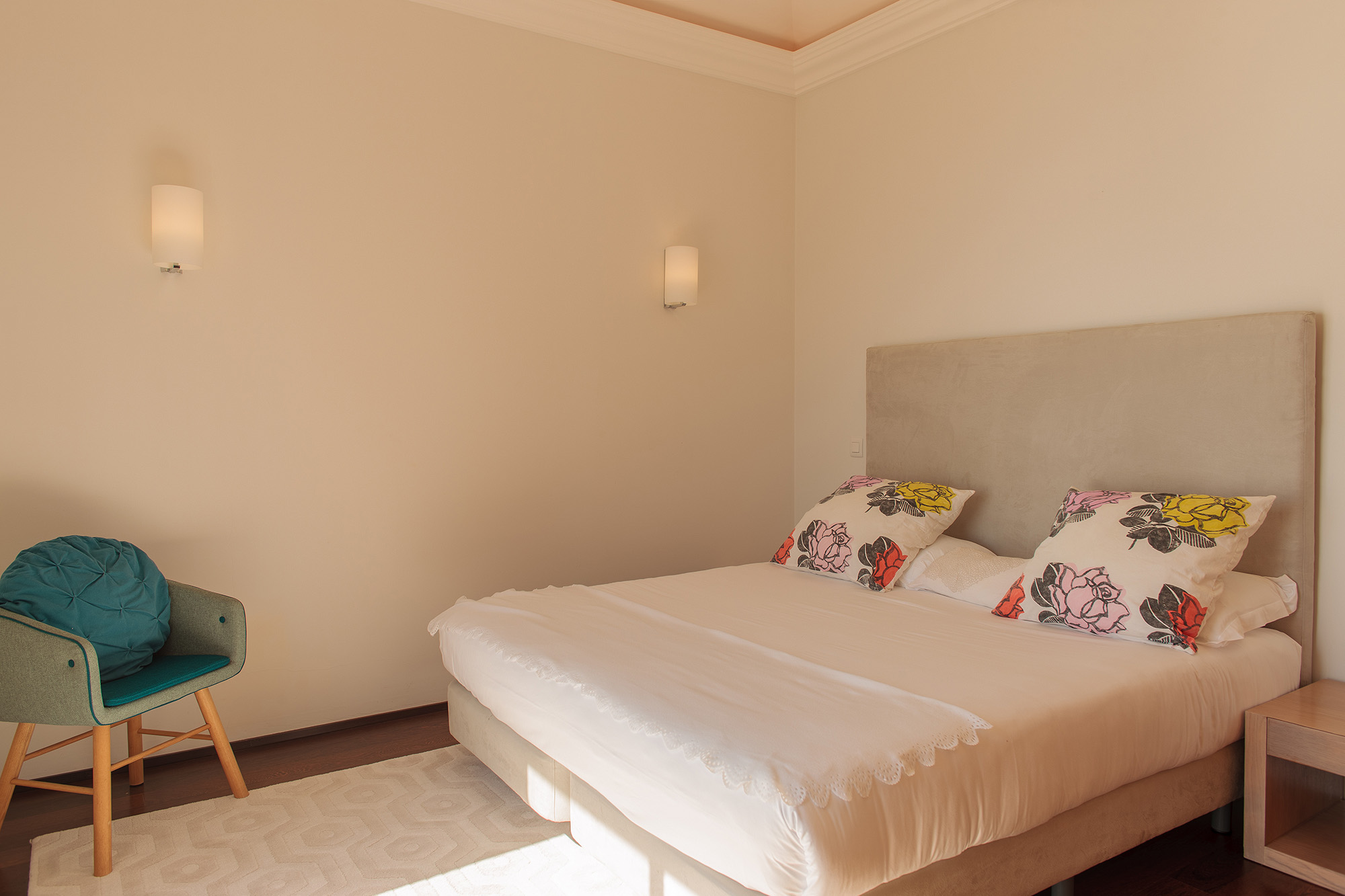 Master bedroom with double bed, colorful floral pillows, teal armchair, wall sconces, and warm morning light, Palheiro Village villa, Madeira