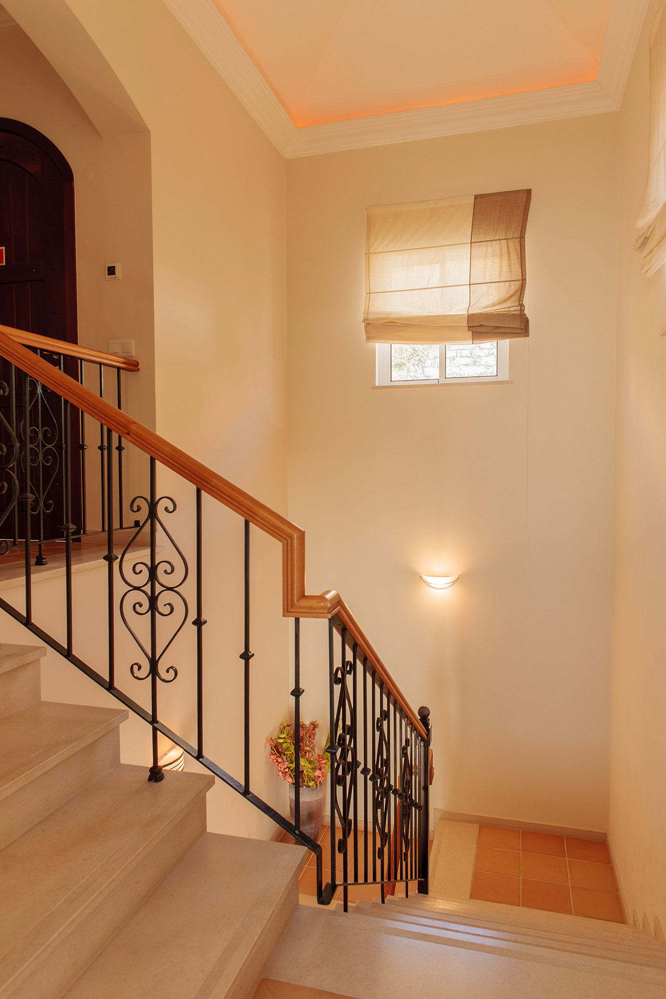 Interior staircase with ornamental black wrought-iron railings, cream walls, light wood handrail, and soft natural light from a landing window, Palheiro Village villa, Madeira