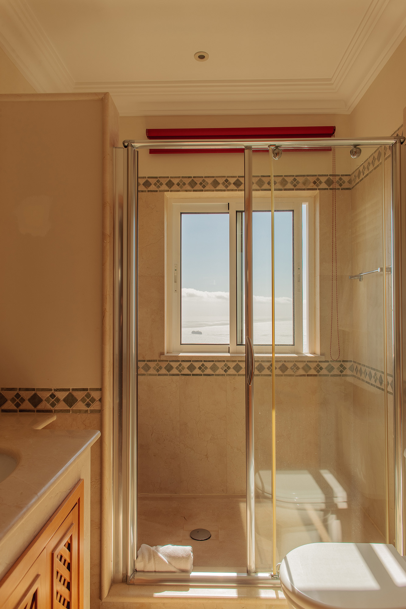 Bathroom in warm beige marble with a glass shower and a small window looking straight out over the sea, Palheiro Village villa, Madeira