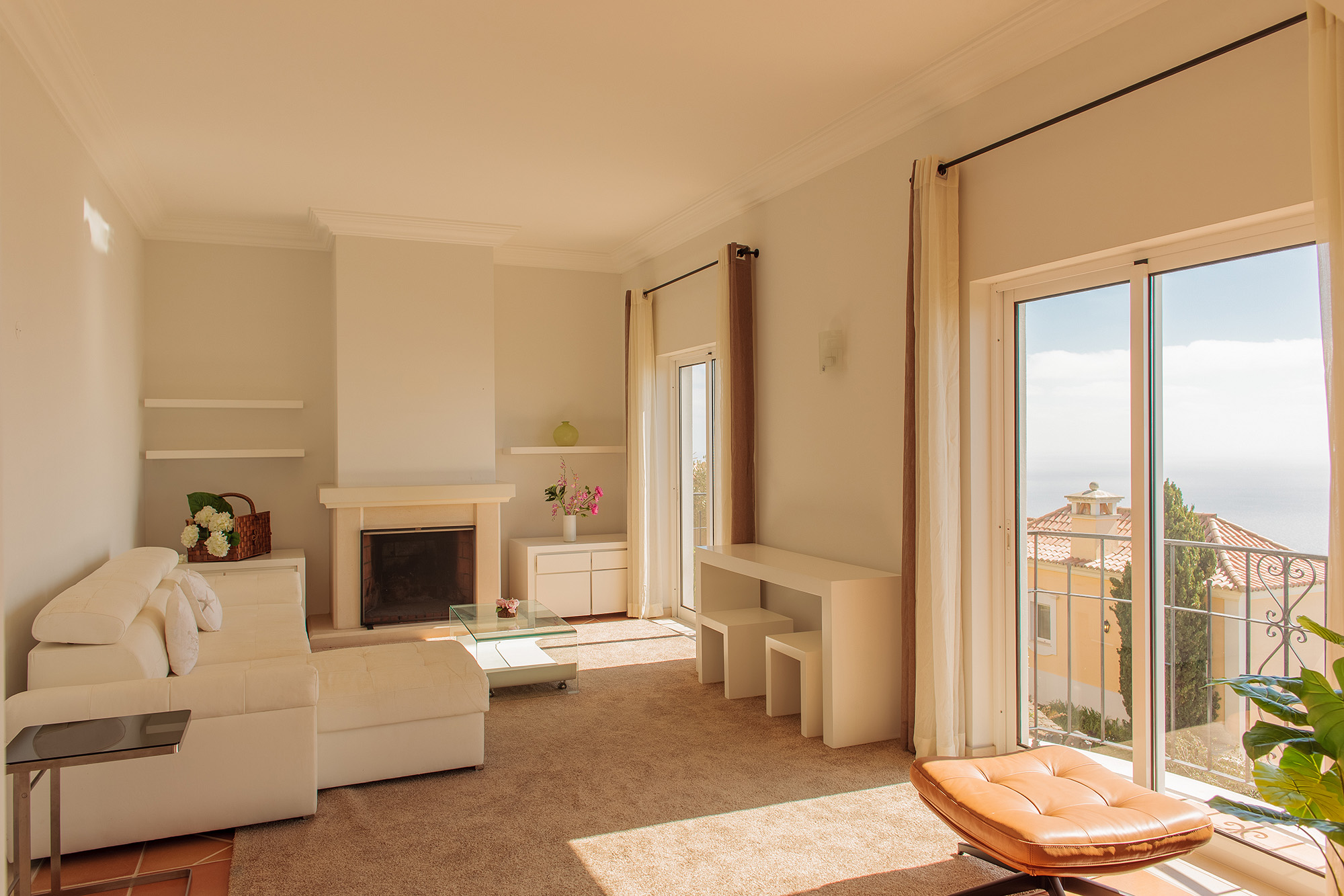 Living room with cream upholstered seating, stone fireplace, natural wood shelving, and glass doors open to terrace with panoramic views, Palheiro Village villa, Madeira