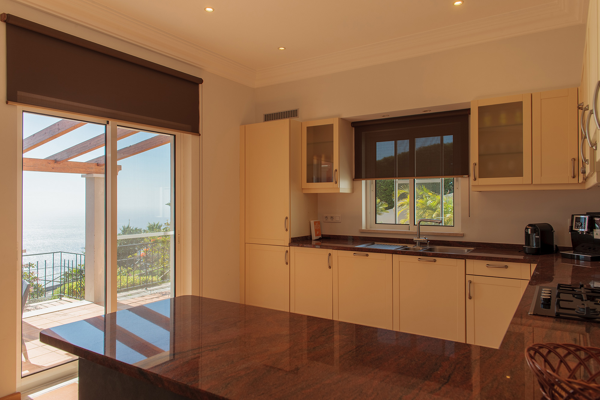 Kitchen with dark granite island, cream cabinets, and sliding glass door opening to pergola terrace with Atlantic ocean view, Palheiro Village villa, Madeira