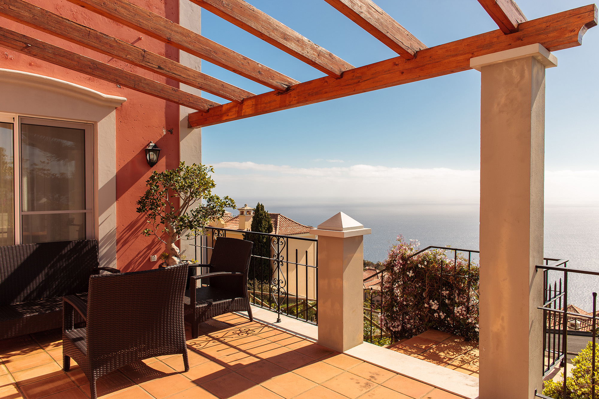 Terrace with wooden pergola, terracotta tiles, wicker chairs, iron railing, and unobstructed Atlantic ocean view over Funchal Bay, Palheiro Village villa, Madeira