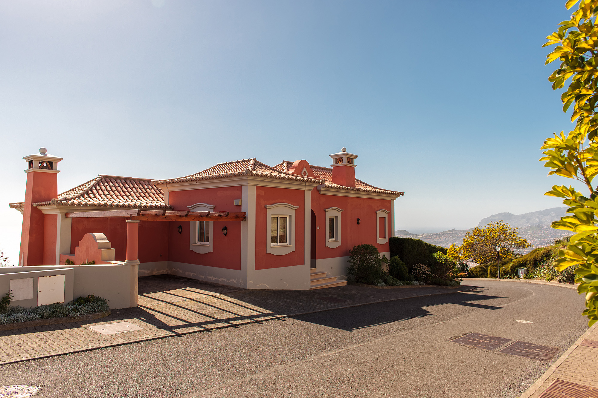 Full exterior view of a Mediterranean villa with red-orange stucco walls, clay barrel tile roof, cream pillars and arched windows, manicured grounds and blue sky, Palheiro Village, Madeira