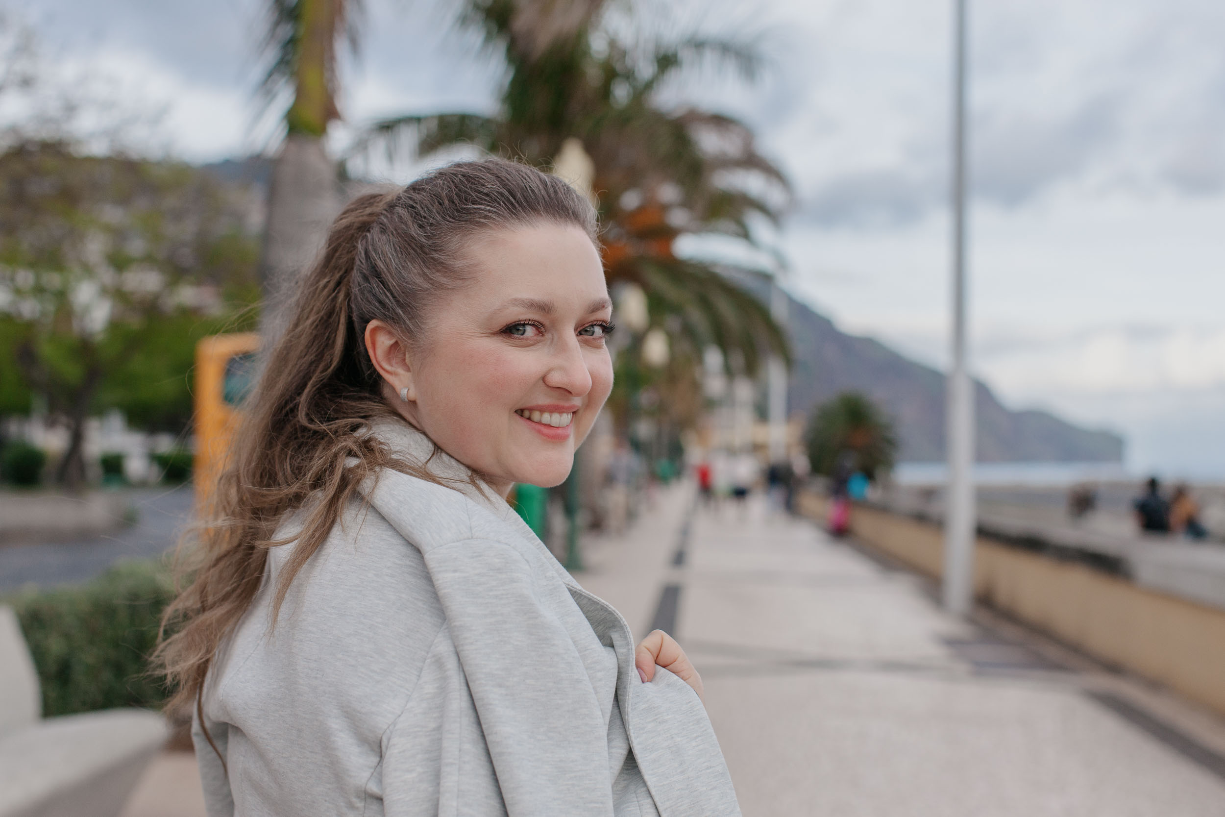 Woman on Funchal promenade looking back over her shoulder at camera, Funchal skyline and hills visible behind, Madeira