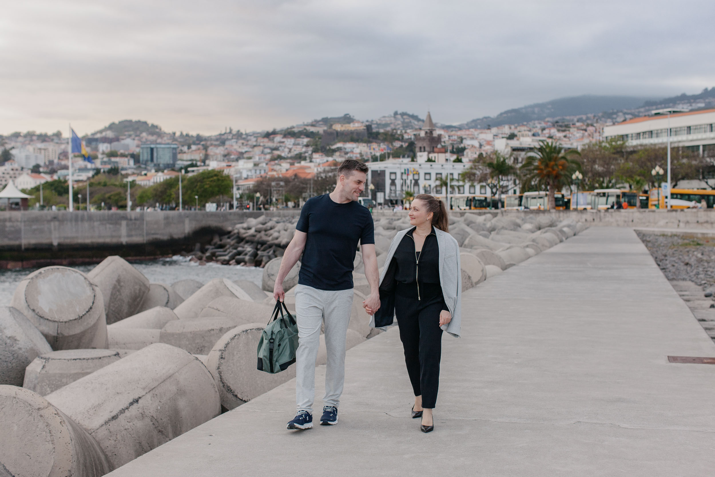 Couple walking along Funchal breakwater pier, Funchal skyline and harbor spread behind them, wide shot, Madeira