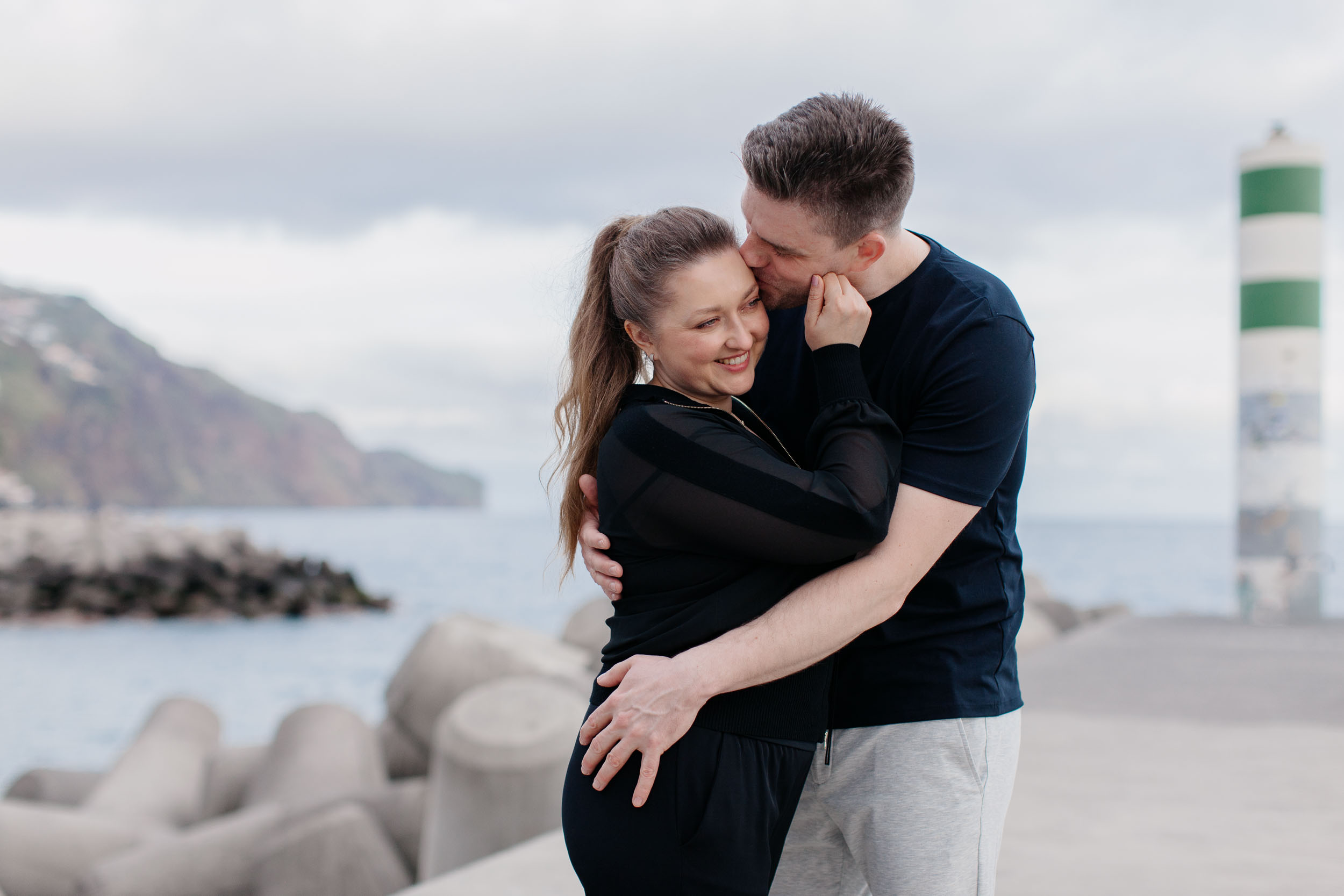 Man kissing woman's temple at Funchal lighthouse, she eyes closed and smiling, cliffs and ocean behind, Madeira