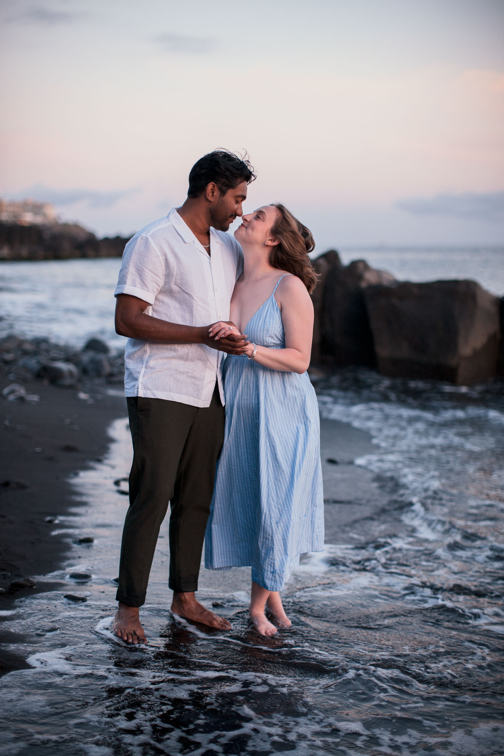 Couple standing barefoot in ocean shallows at dusk, about to kiss, Praia Formosa, Madeira