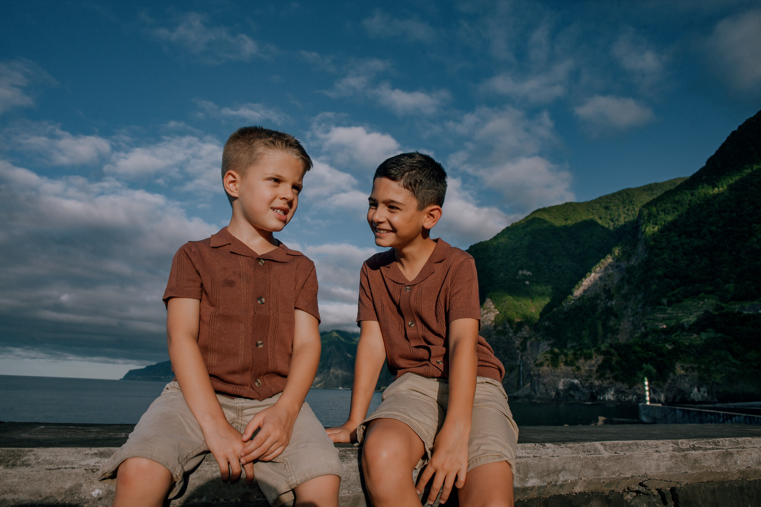 The boys side by side on the seawall, cliffs across the water, Seixal, Madeira
