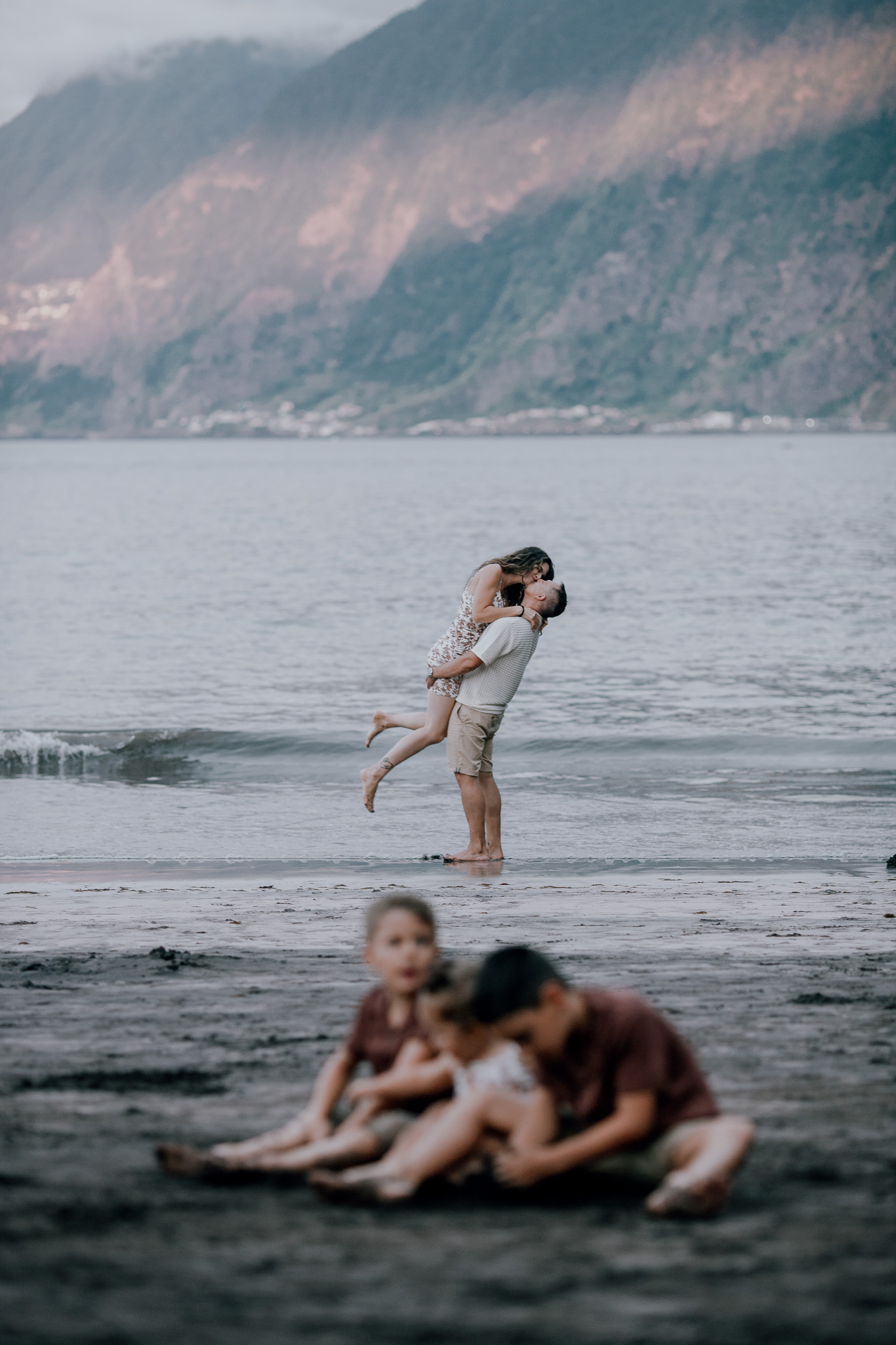 He spins her in the air on the dark beach, children playing behind, Seixal, Madeira