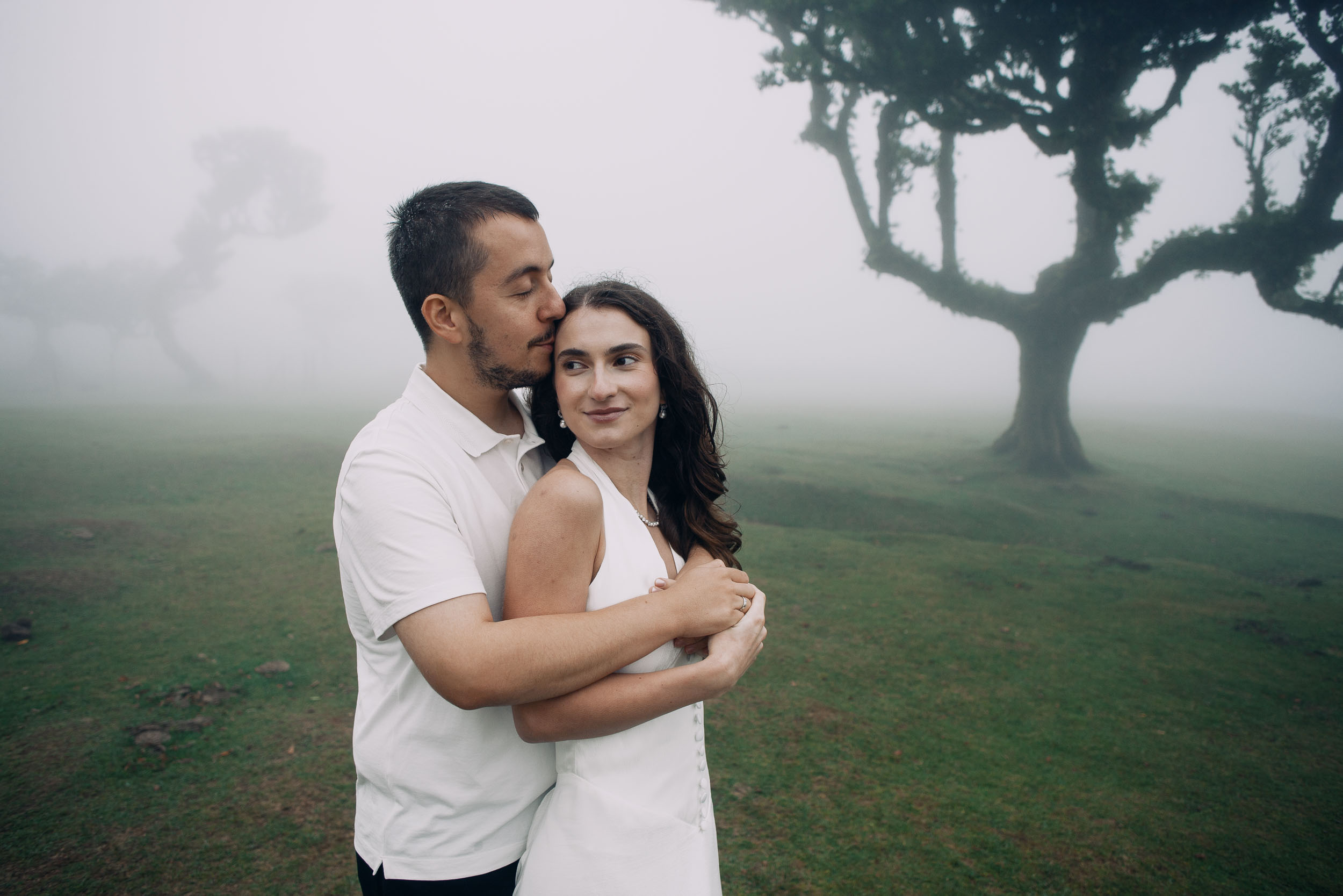 Man holding his wife from behind while she looks off to the side, laurel grove in mist, Fanal Forest, Madeira