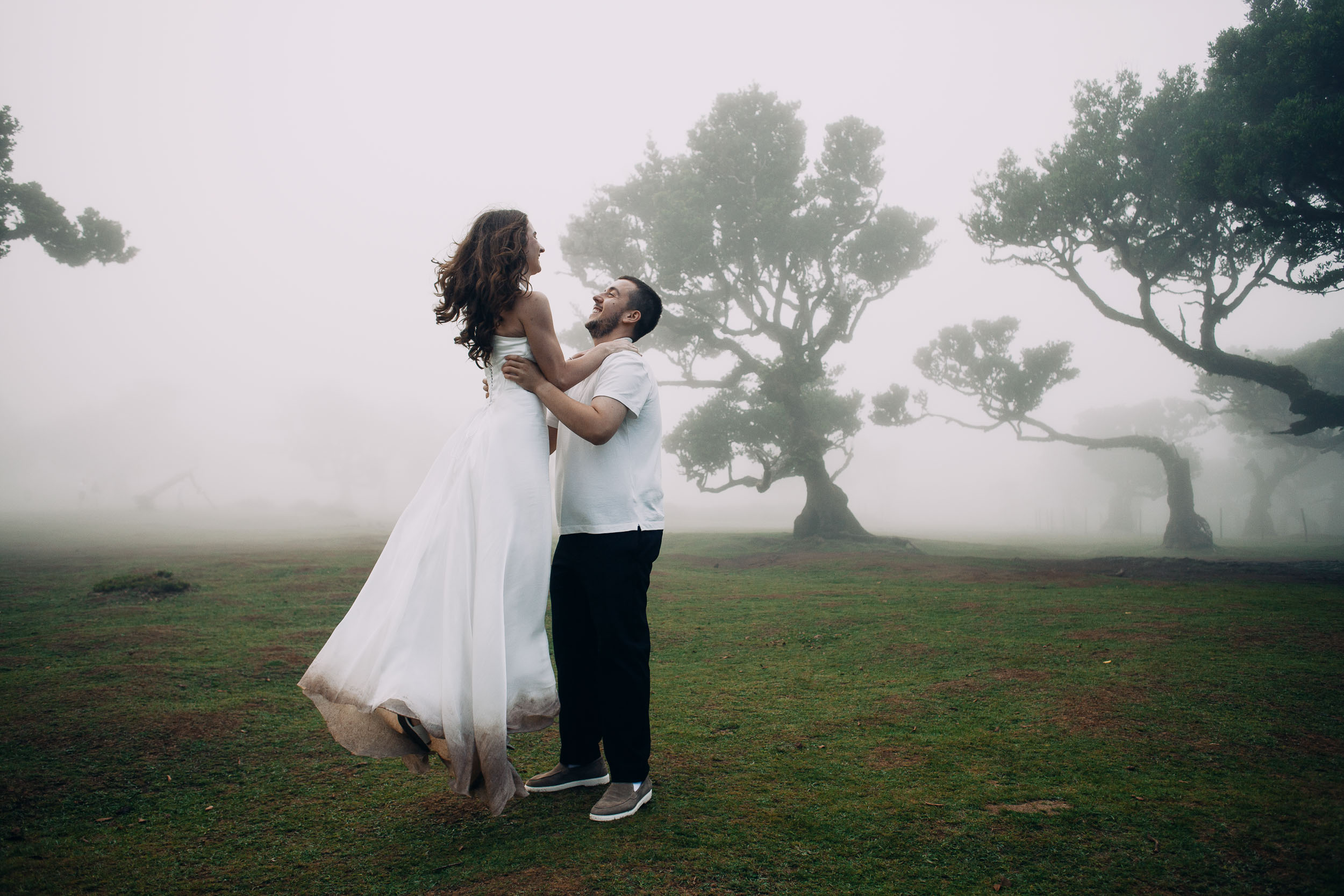 Man lifting his wife off the ground, both laughing, laurel trees behind, Fanal Forest, Madeira