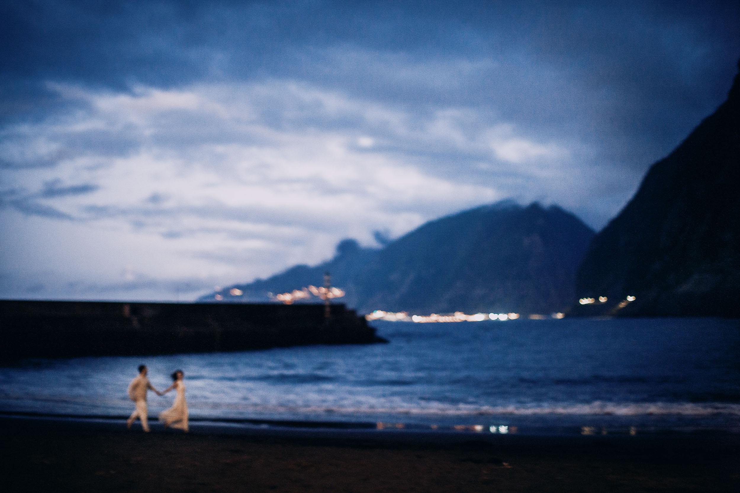 Wide blue hour frame of the couple on the black sand, breakwater and village lights across the bay, Seixal Beach, Madeira