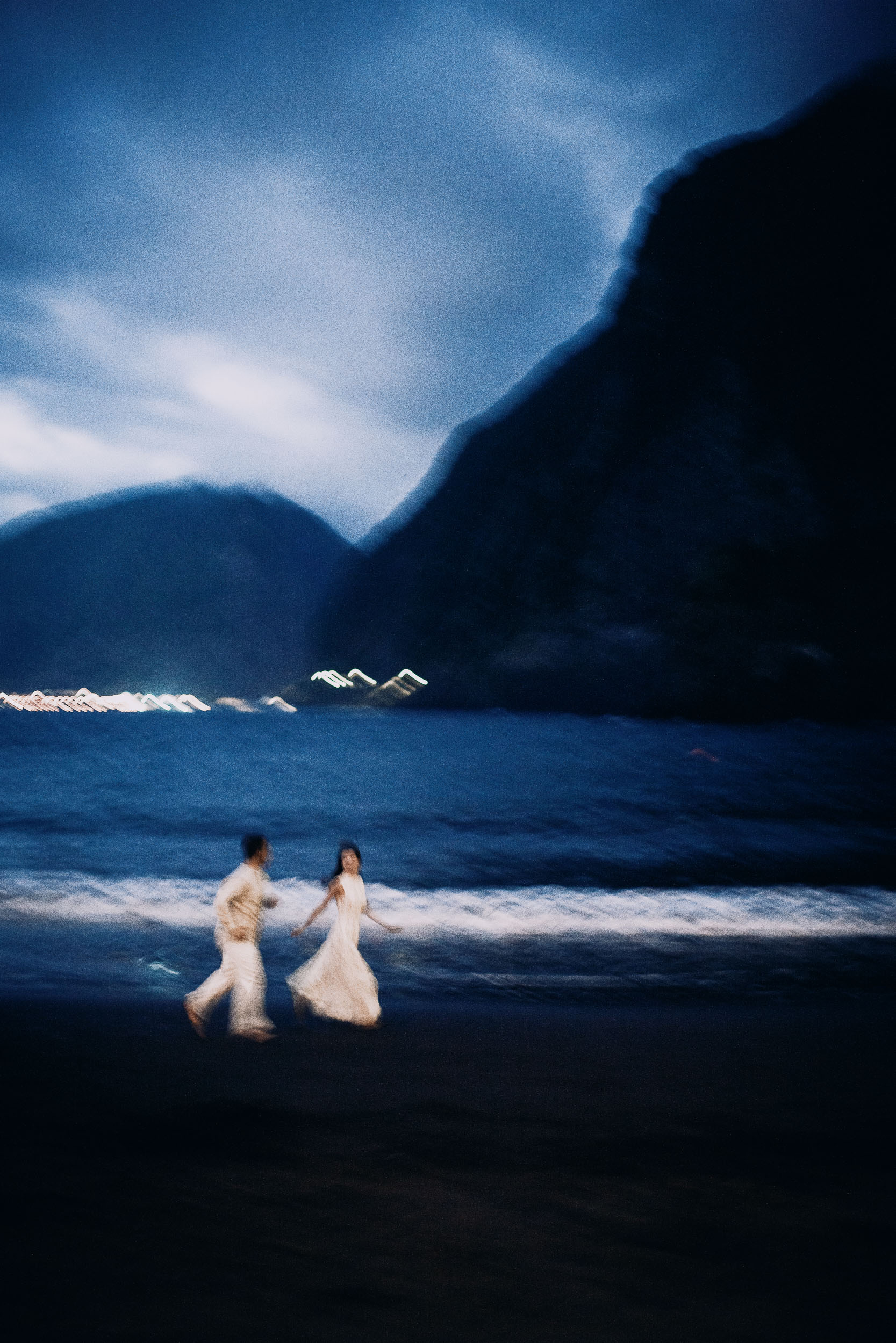 Couple running through the shallows half an hour after sunset, coast road lights stretched across the bay, Seixal Beach, Madeira