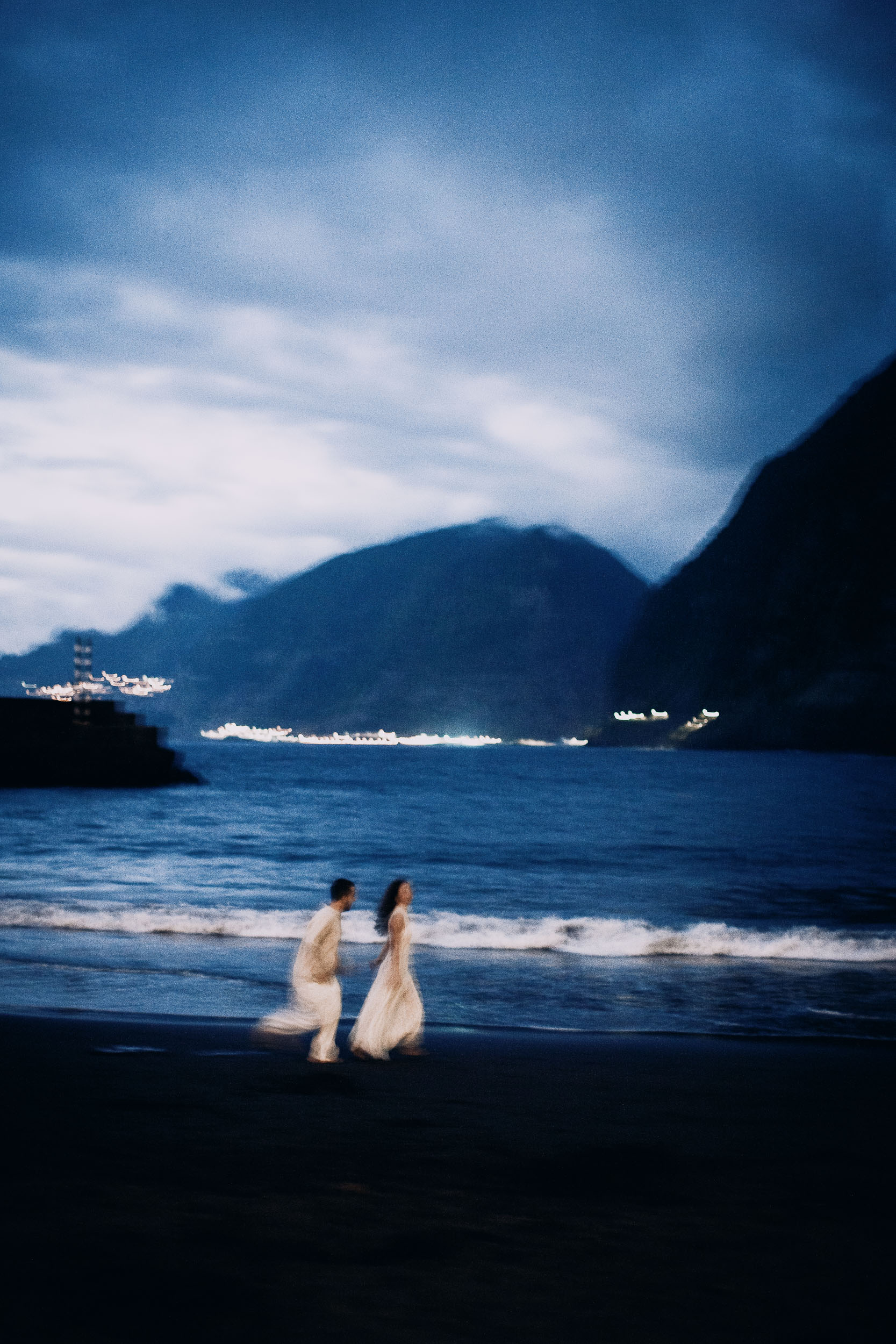Couple walking at the water's edge, village lights on the cliffs behind them, Seixal Beach, Madeira