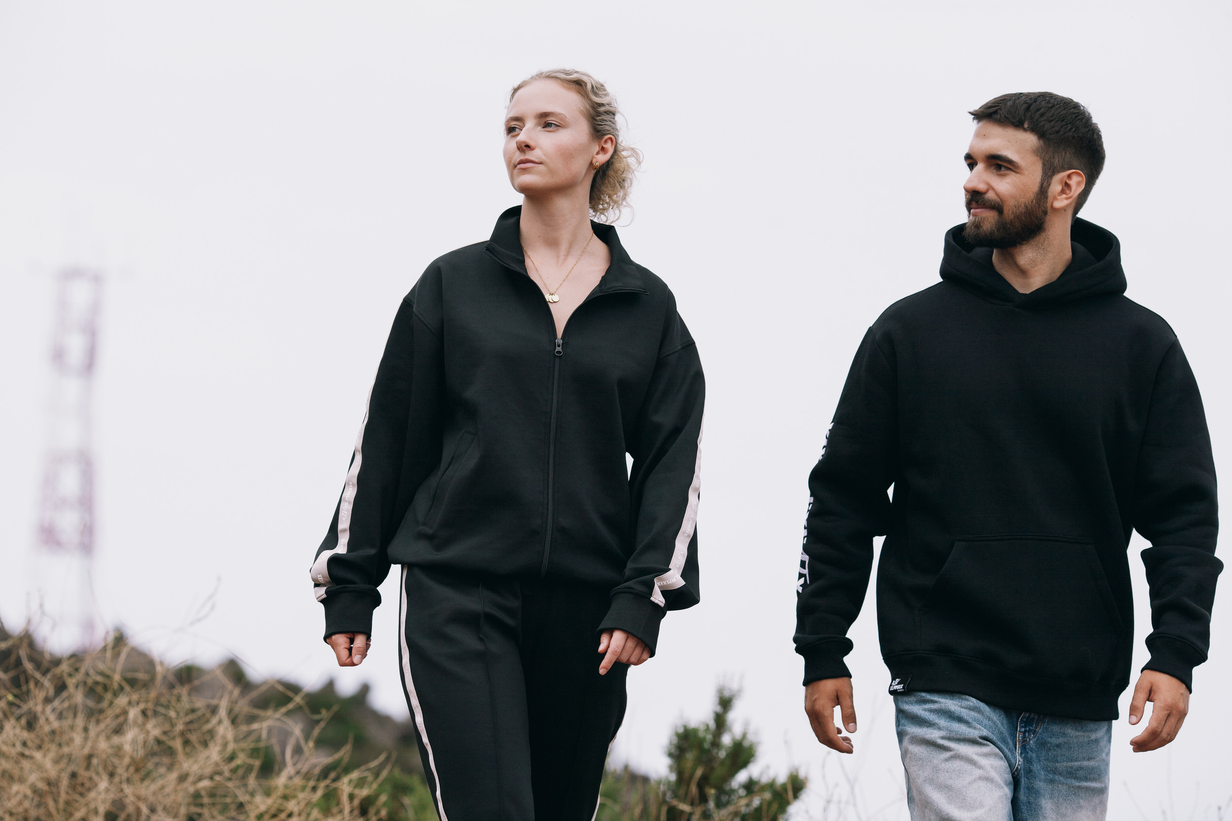 Wider frame of the same pair walking through dry grass in black Elotrix outerwear, Madeira highlands