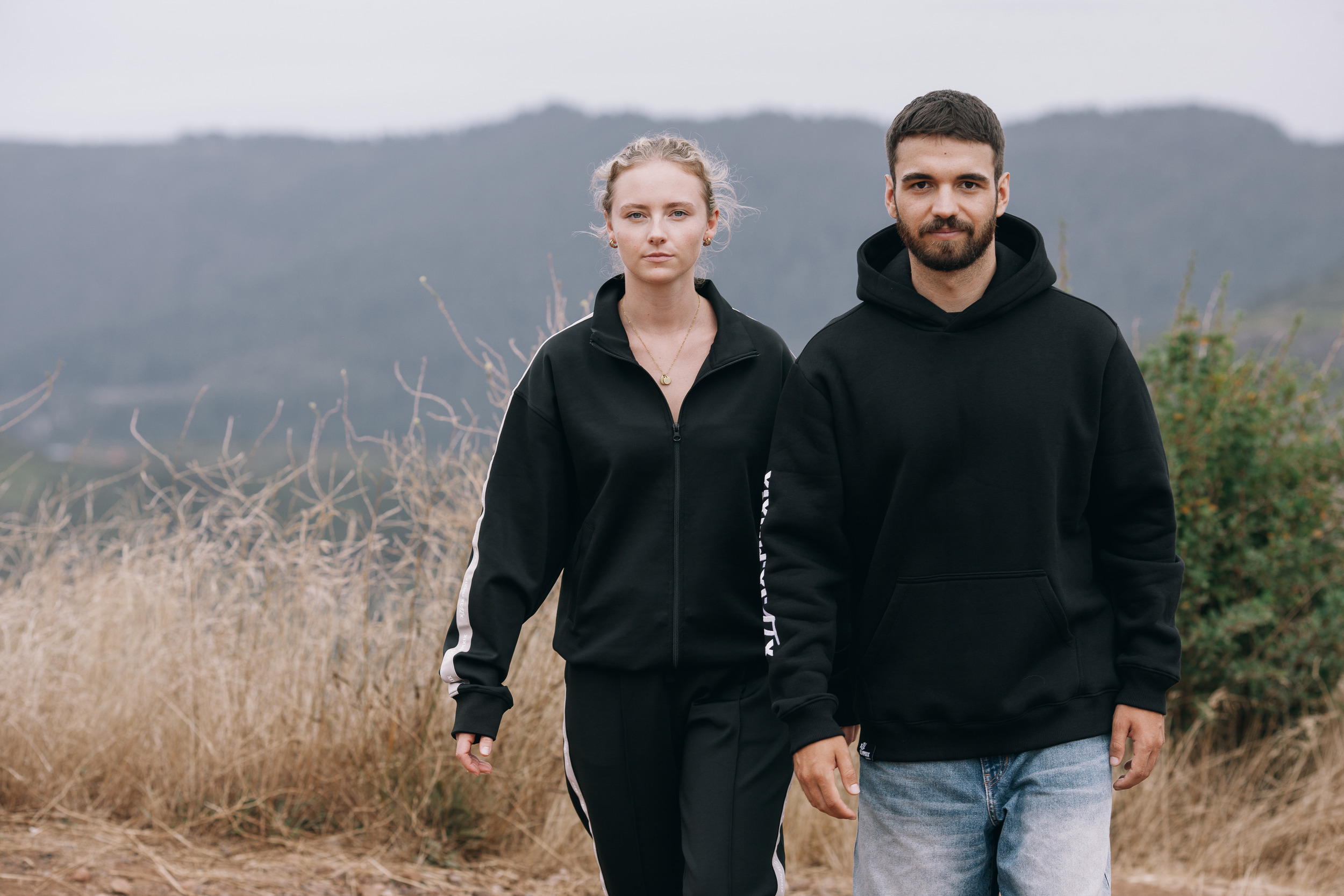 Model pair standing still in the dry grass, mountain ridges flattened by Saharan dust behind them, Madeira