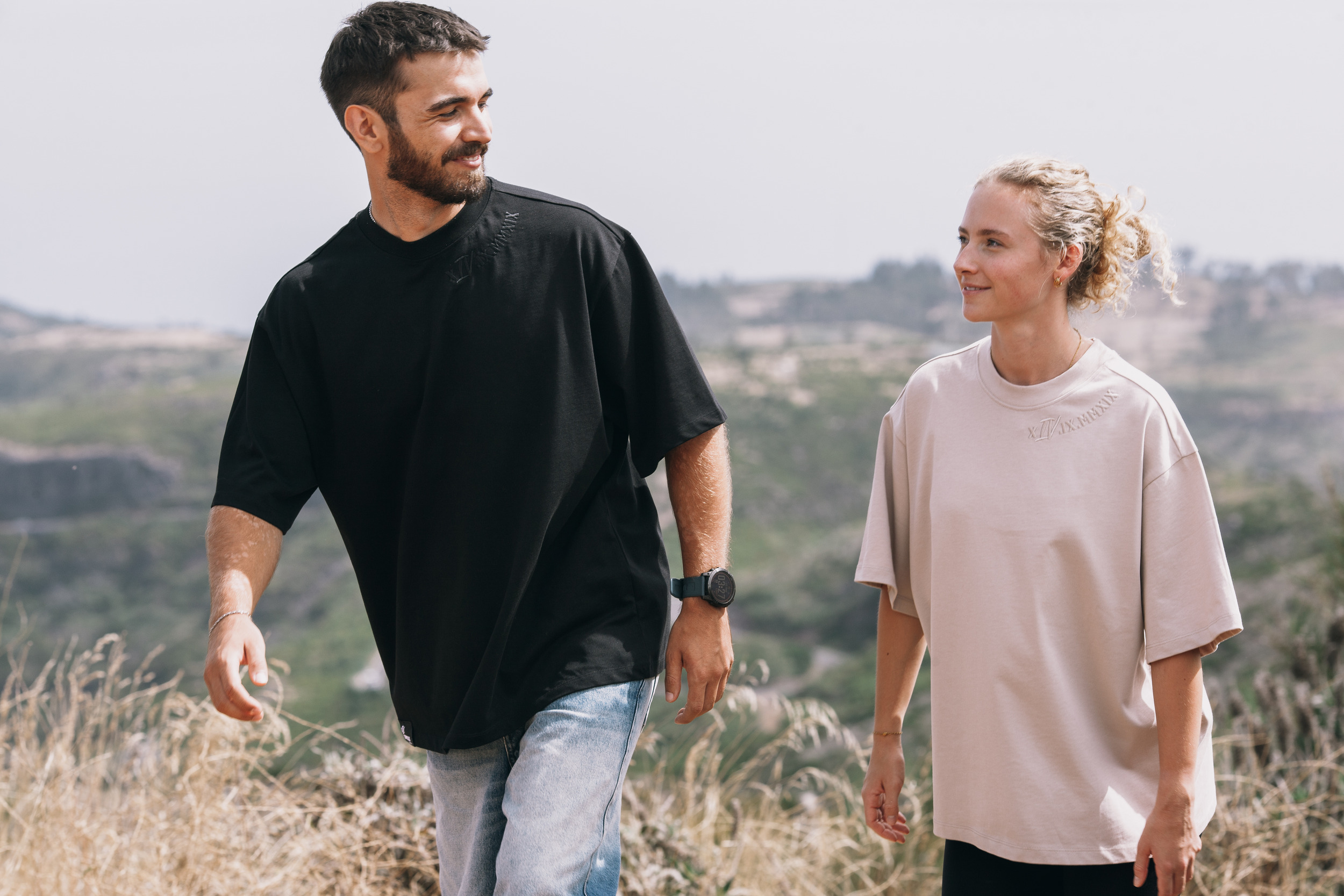 Man in an oversized black tee and woman in an oversized beige tee walking along the ridge, Madeira