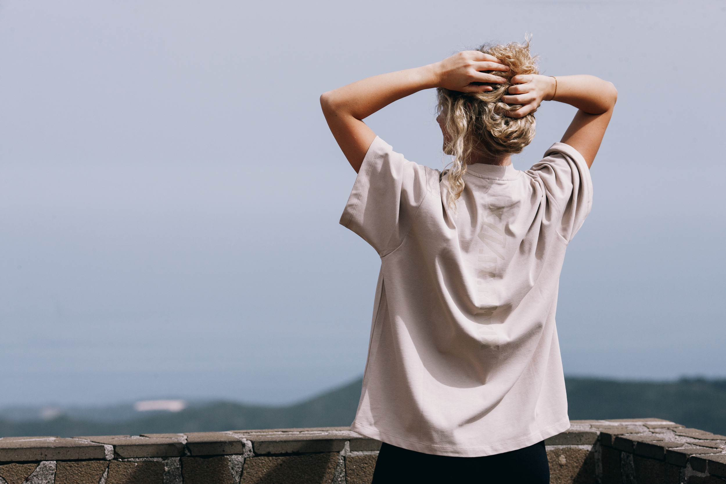 Woman seen from behind with both hands in her hair, beige oversized tee against the hazy sea, Madeira