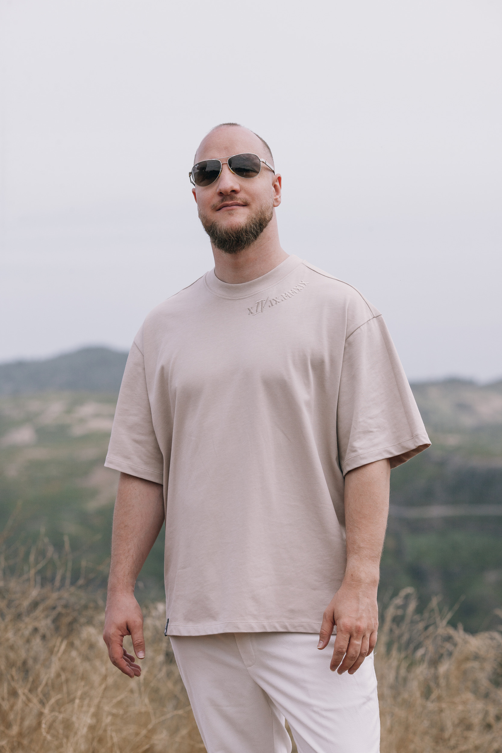 Man in a beige oversized tee and sunglasses standing against the hazy mountains, Madeira