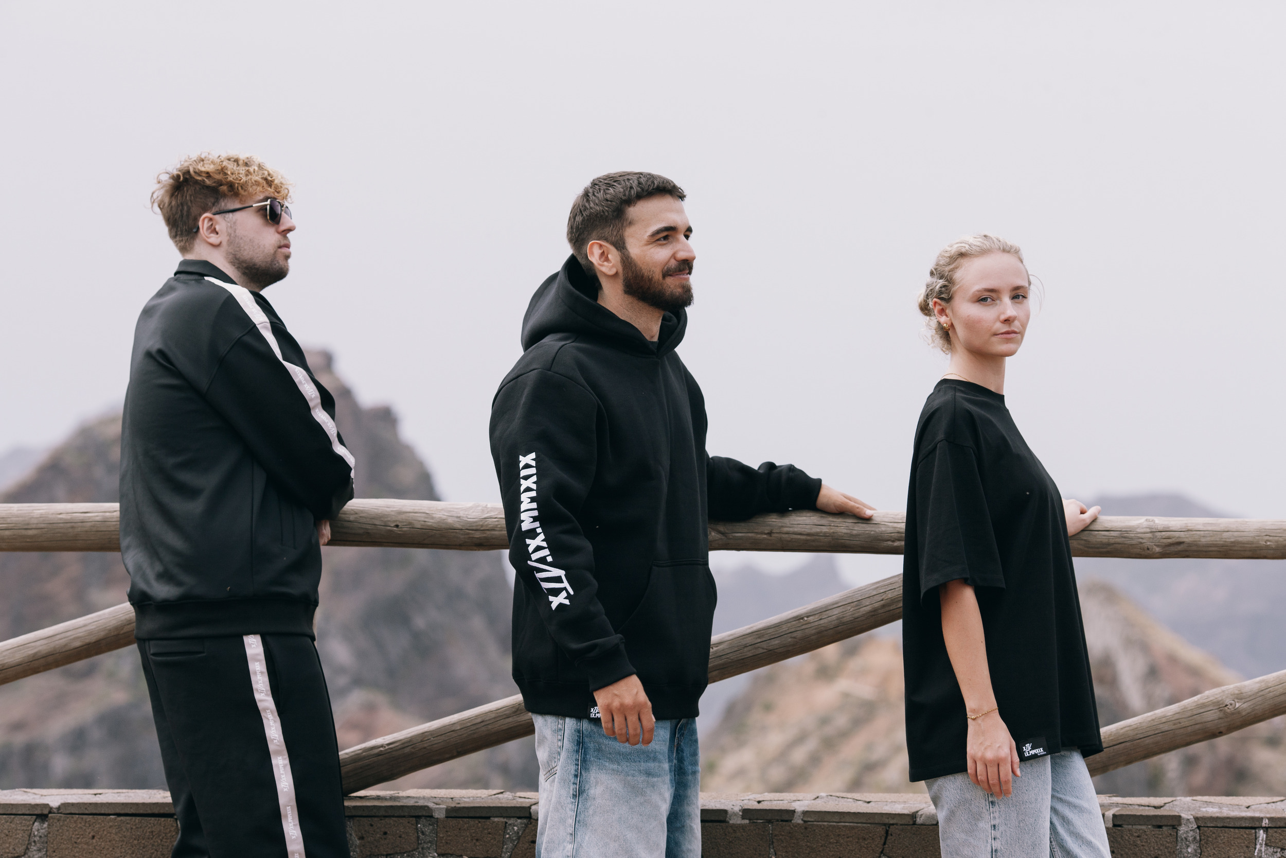 Three models in profile along the railing, mountains flattened by dust behind, Madeira