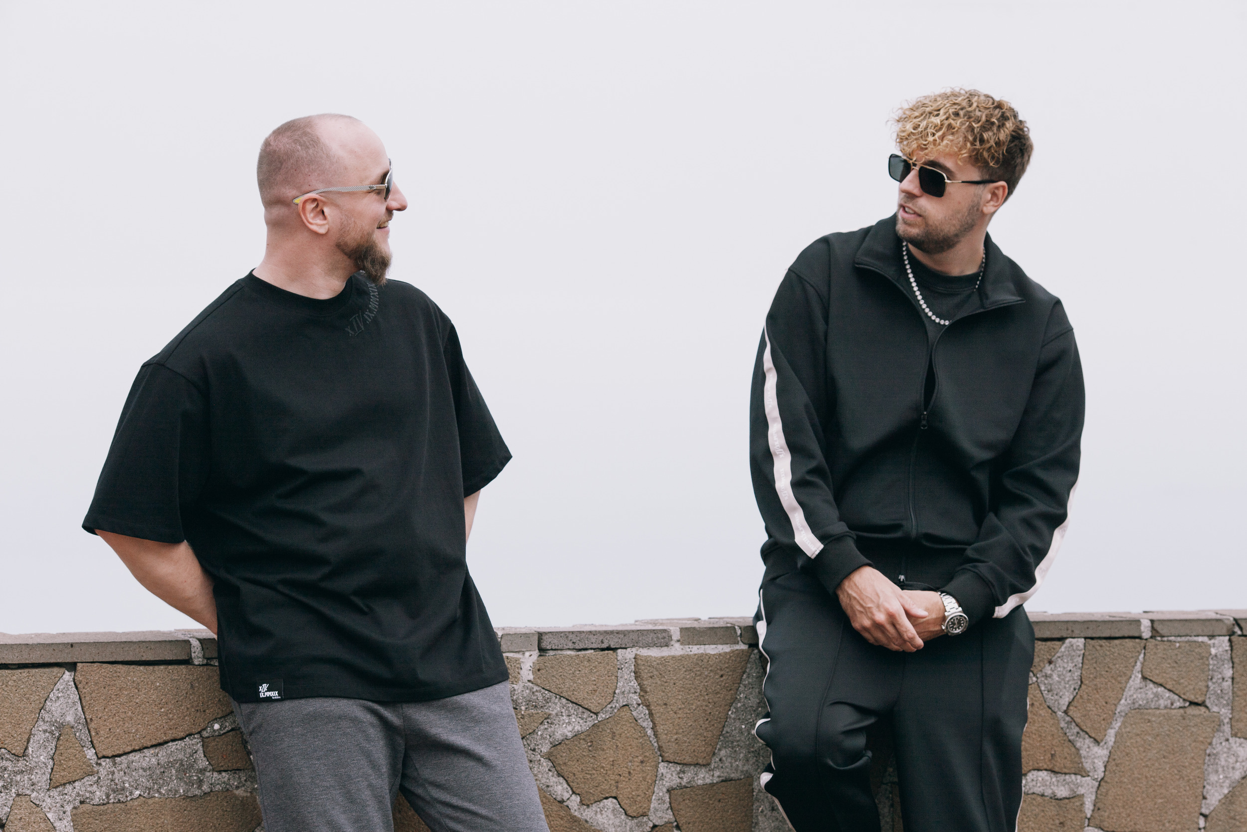 Two men leaning on the stone wall in black tee and track jacket, Madeira