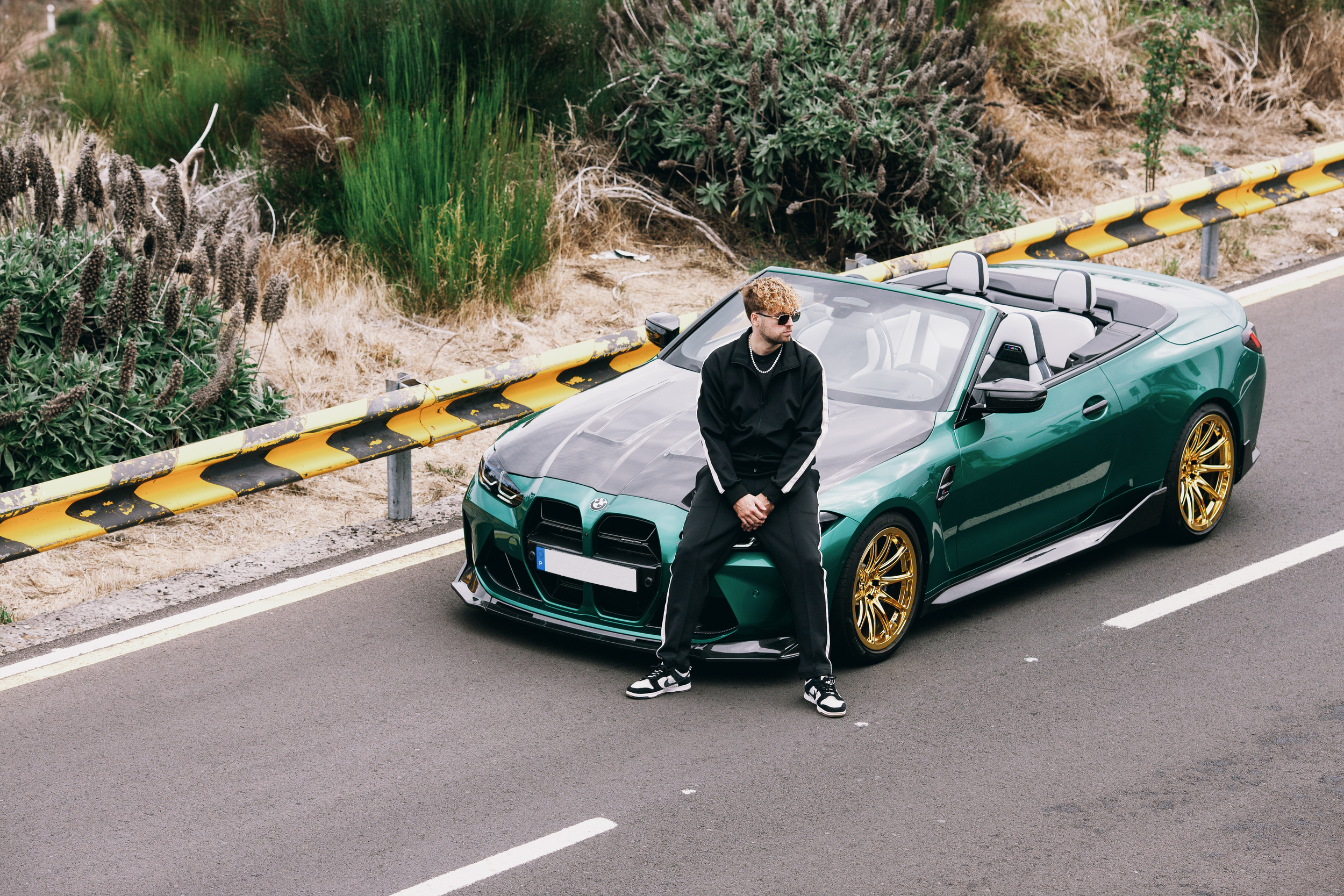 Man in a black track suit sitting on the bonnet of a green convertible on a mountain road, Madeira