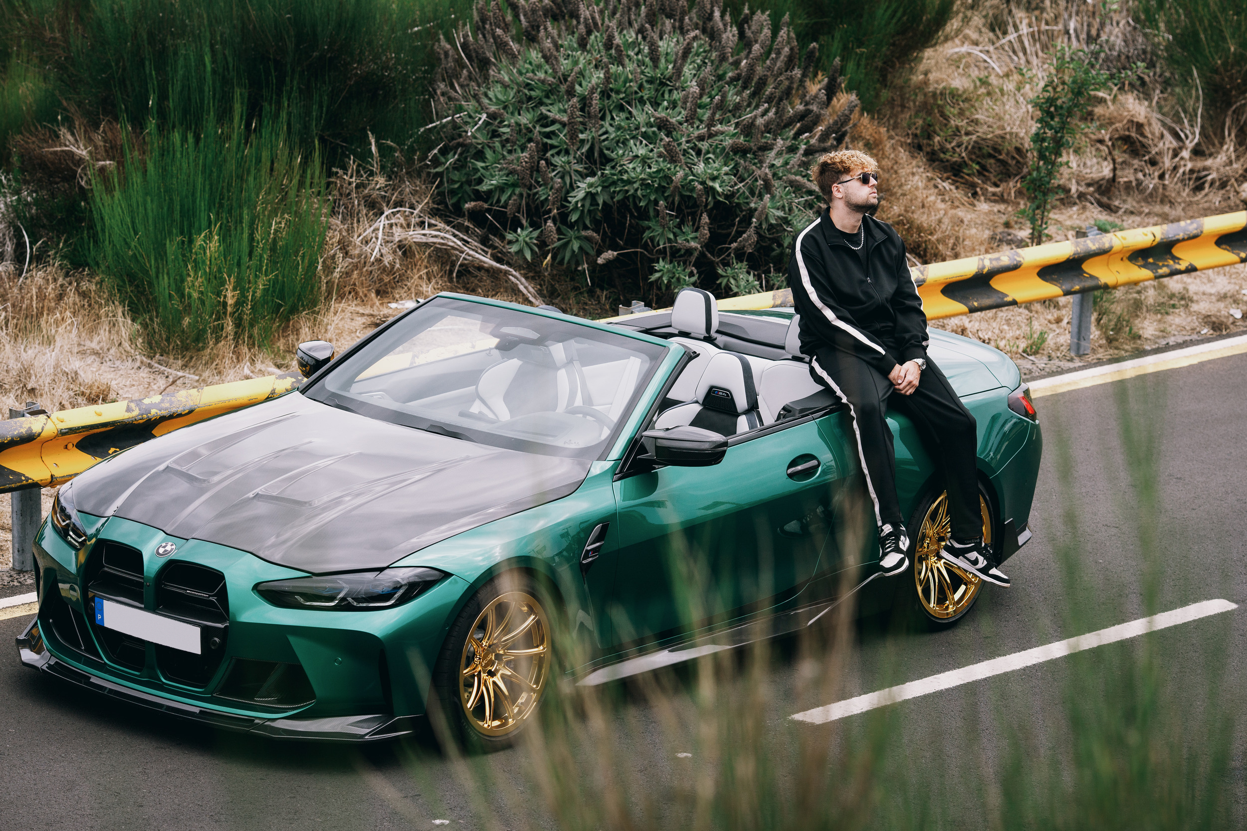Green convertible on the mountain road framed by grass, model on the bonnet, Madeira