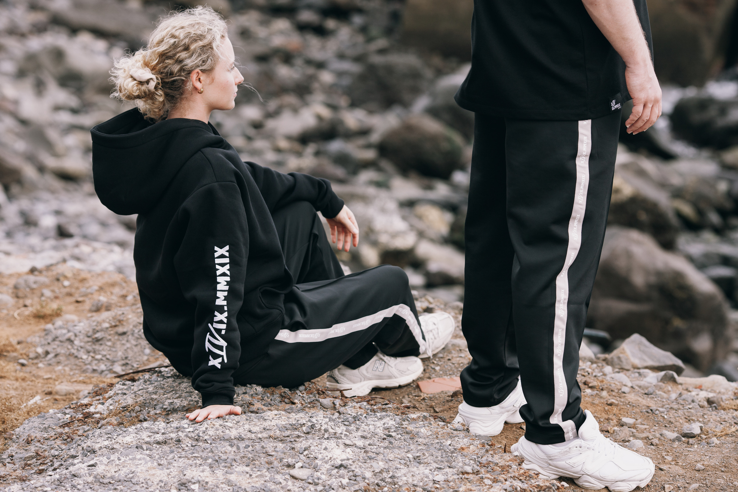Woman sitting on the rocks in a hoodie while a model stands beside her, Madeira