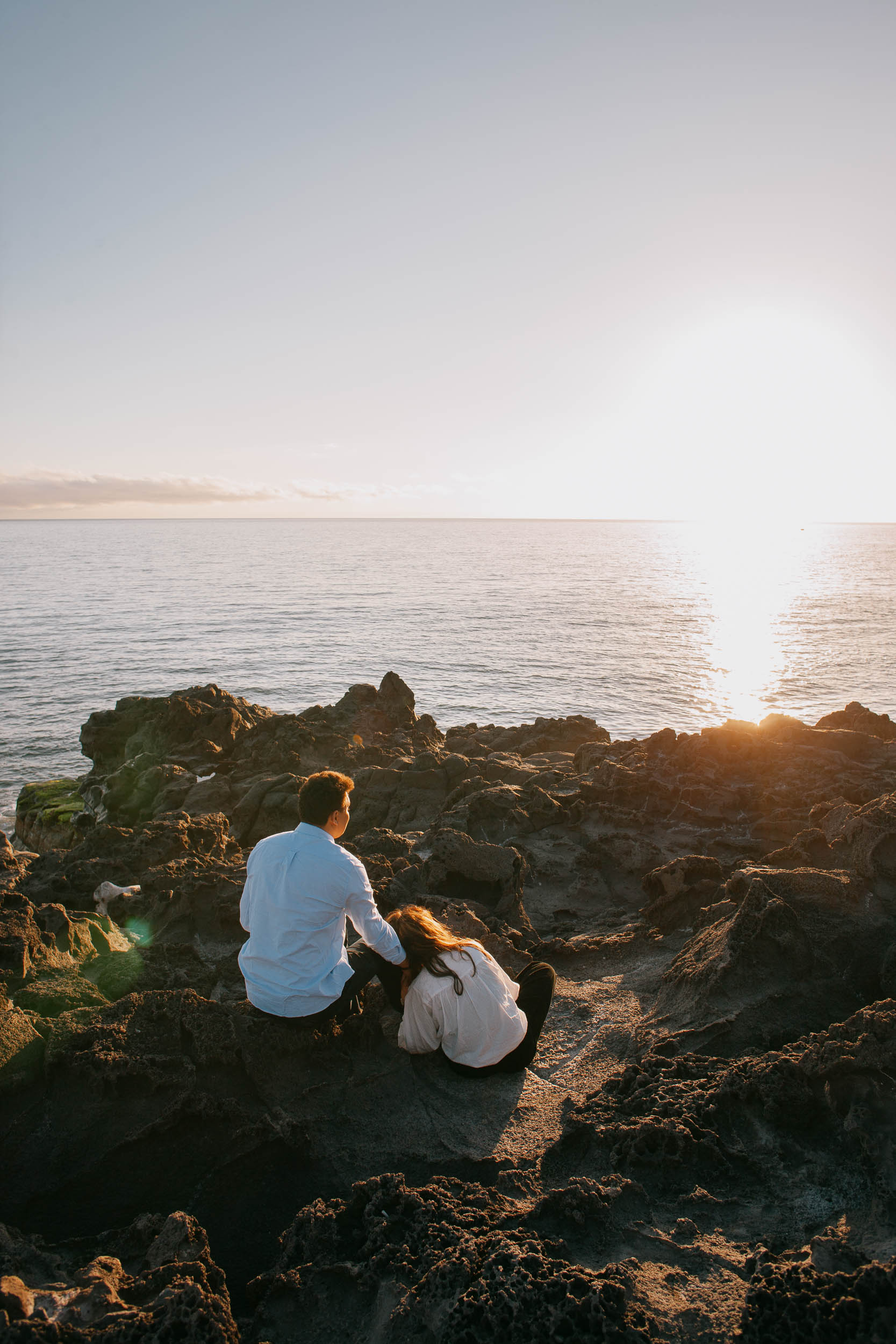 Couple laughing together on volcanic rocks at Praia Formosa, cliffs of Funchal in the background, golden sunset light, Madeira