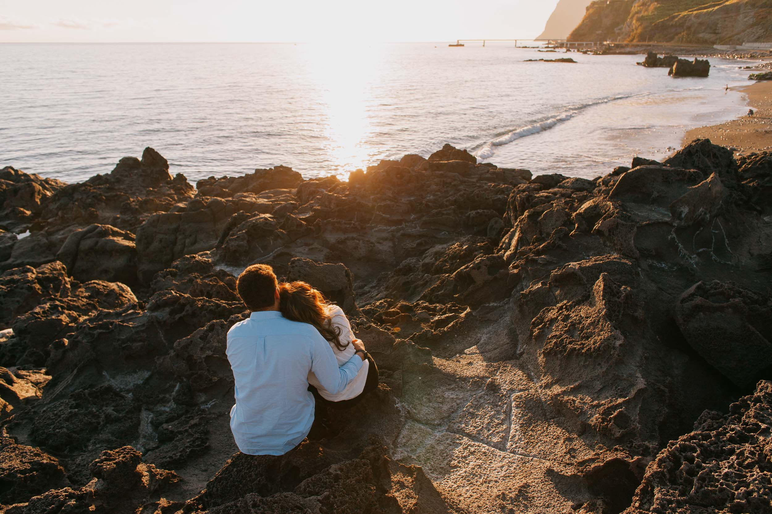 Couple sitting on volcanic rocks watching the sunset at Praia Formosa, backs to camera, Madeira