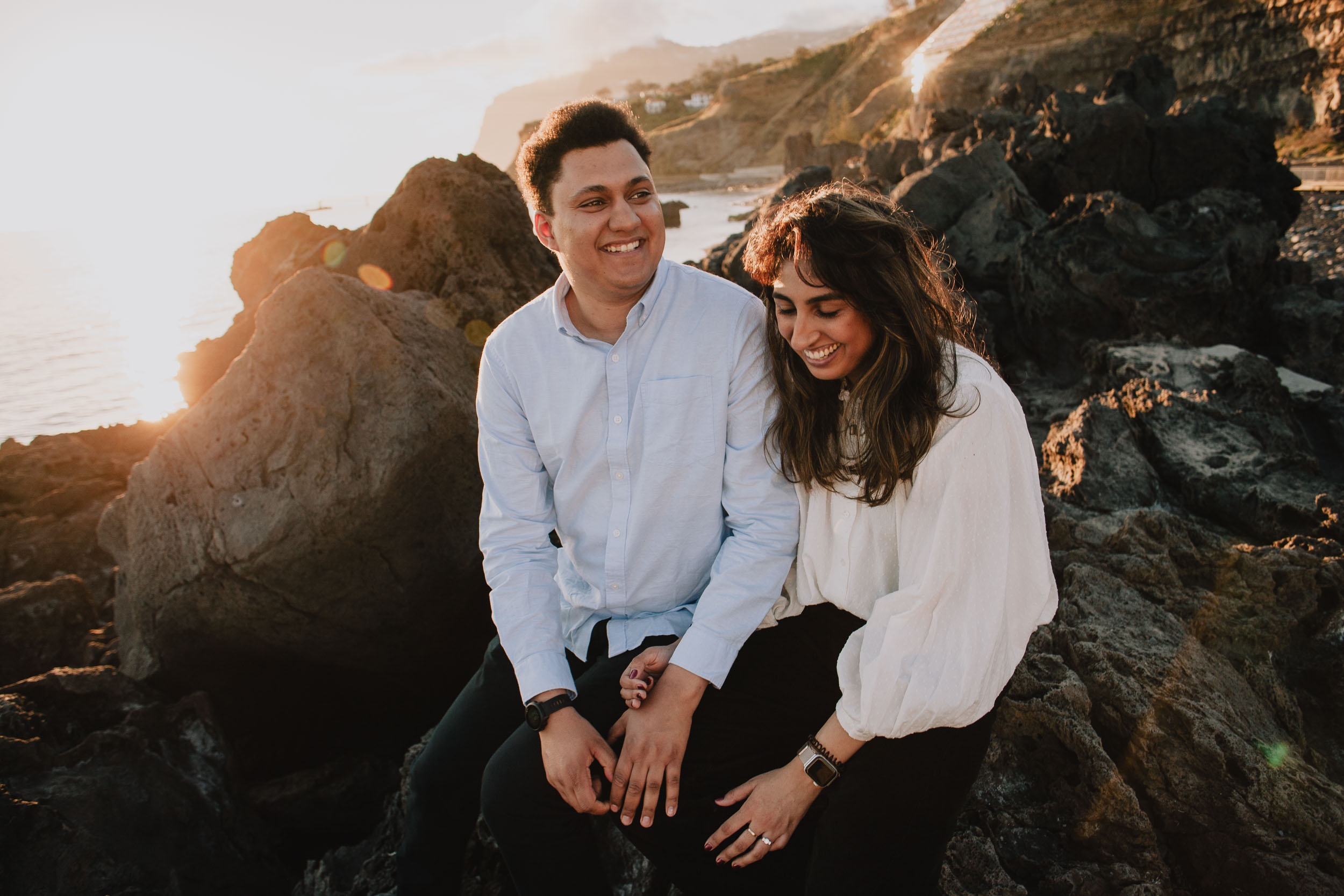 Couple sitting on volcanic rocks at Praia Formosa, wide view of the beach and Atlantic coastline behind them, sunset, Madeira