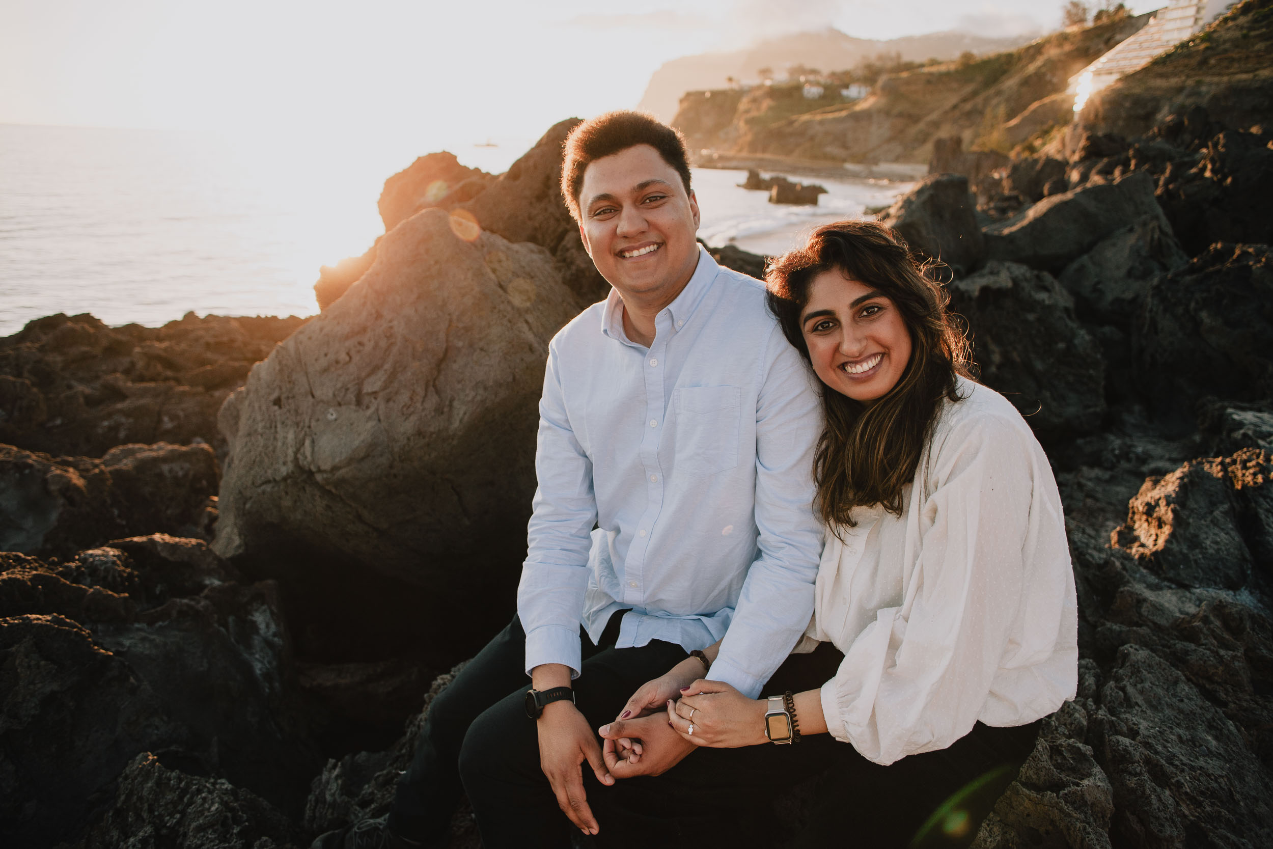 Couple smiling at camera seated on volcanic rocks at Praia Formosa, warm golden sunset light, Funchal cliffs and Atlantic behind, Madeira