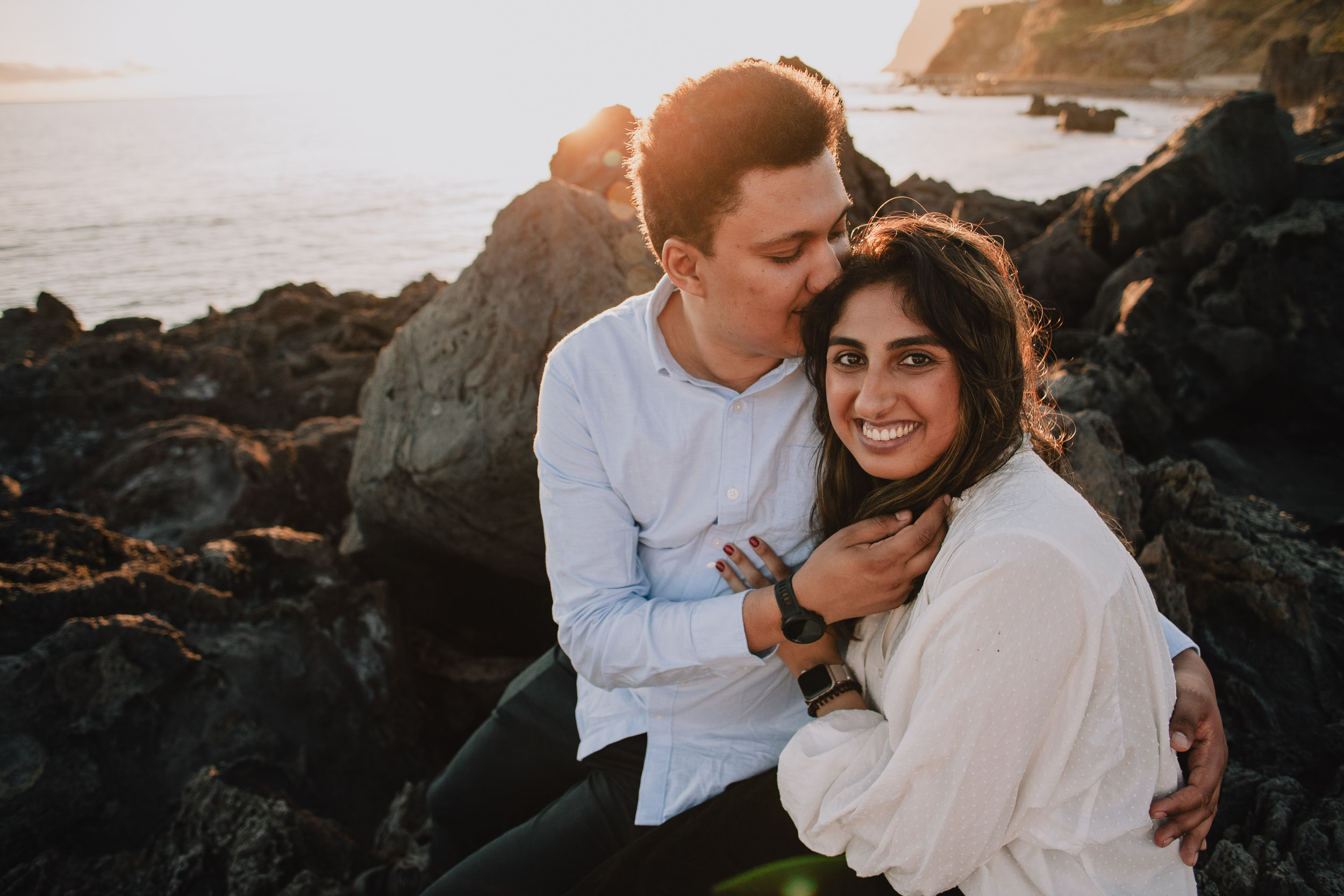 Man kissing woman's temple while she smiles broadly at camera, strong golden sunset light, Funchal cliffs behind, Praia Formosa, Madeira