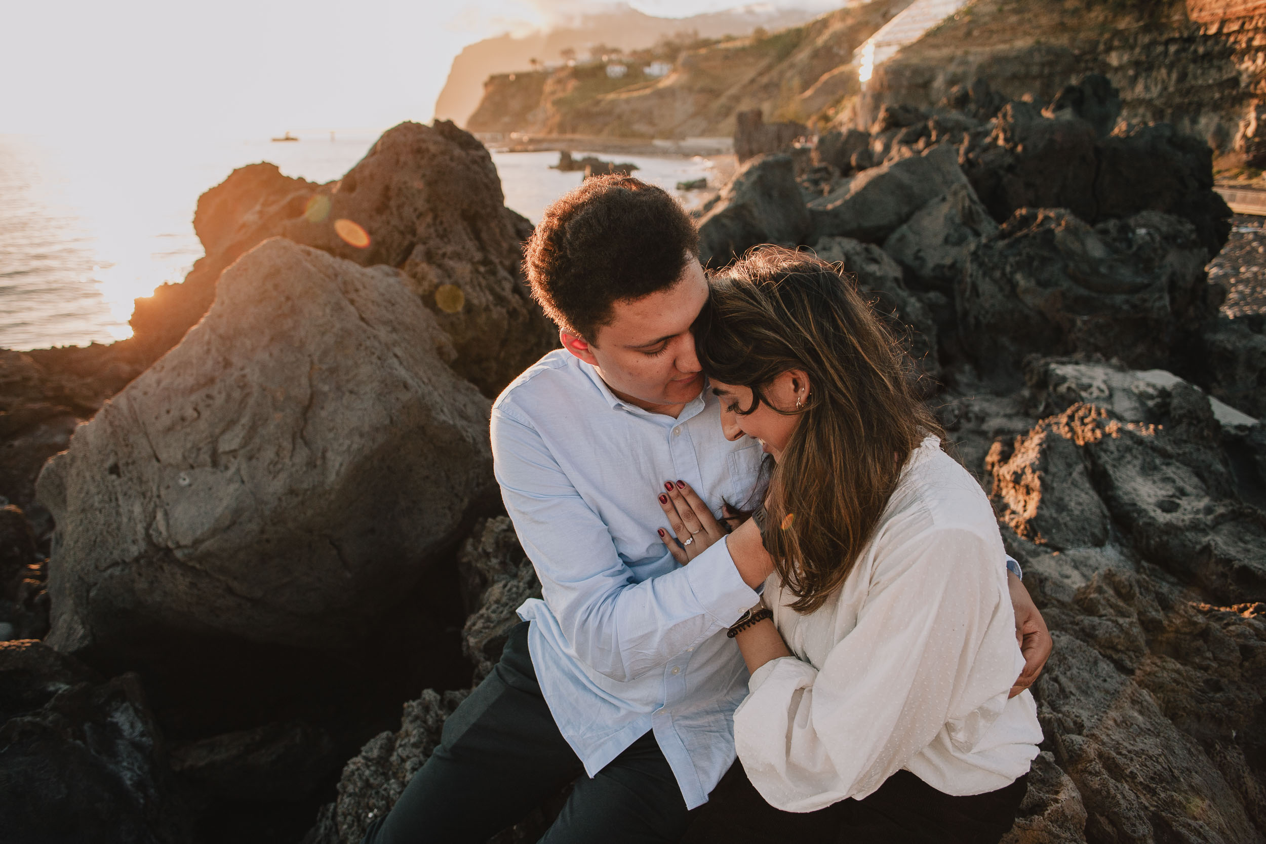 Man holding woman from behind on volcanic rocks, nuzzling her cheek, golden sunset light, Praia Formosa, Madeira