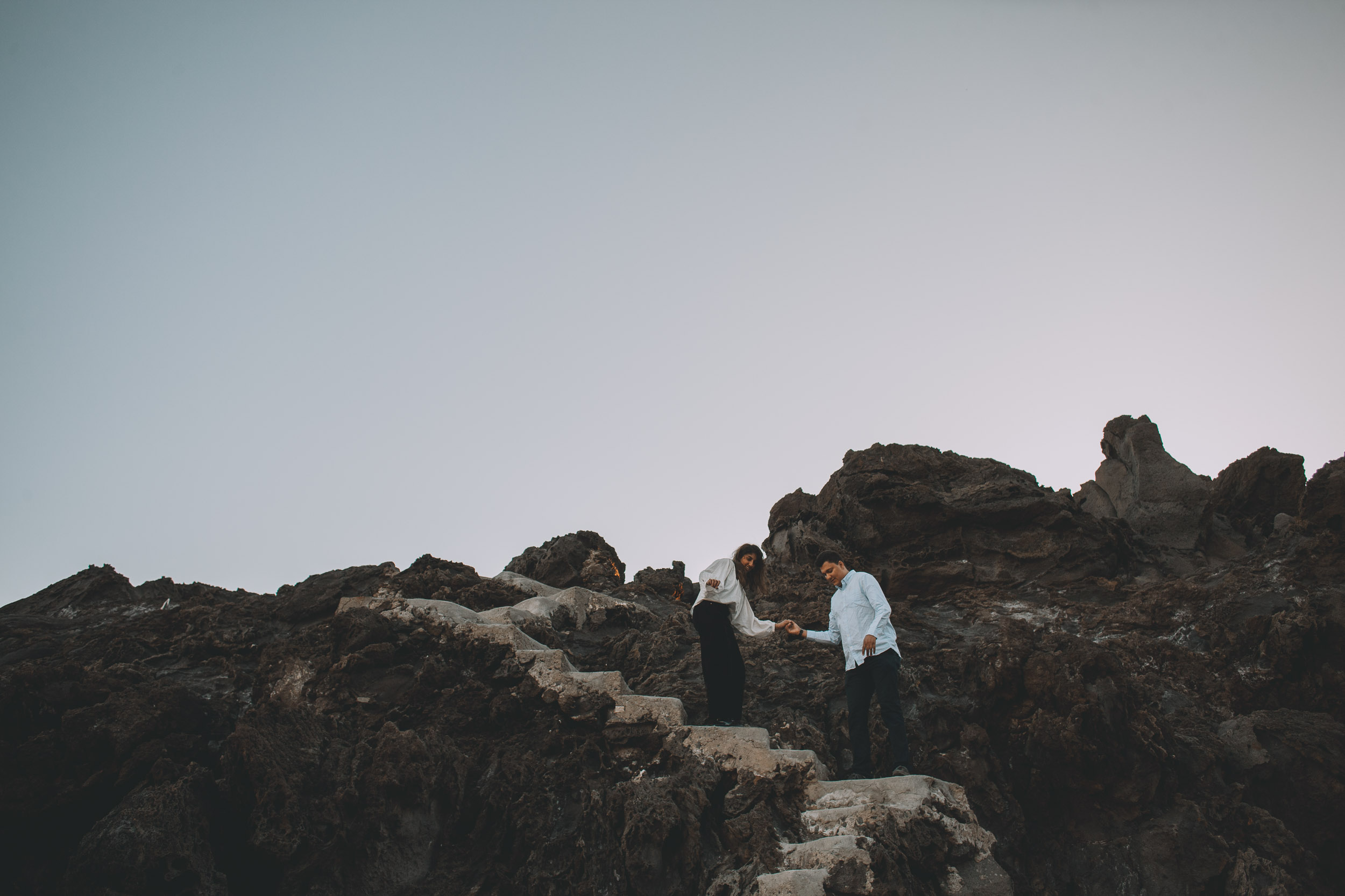Couple helping each other down stone steps cut into volcanic rock at Praia Formosa, wide pale sky above, Madeira