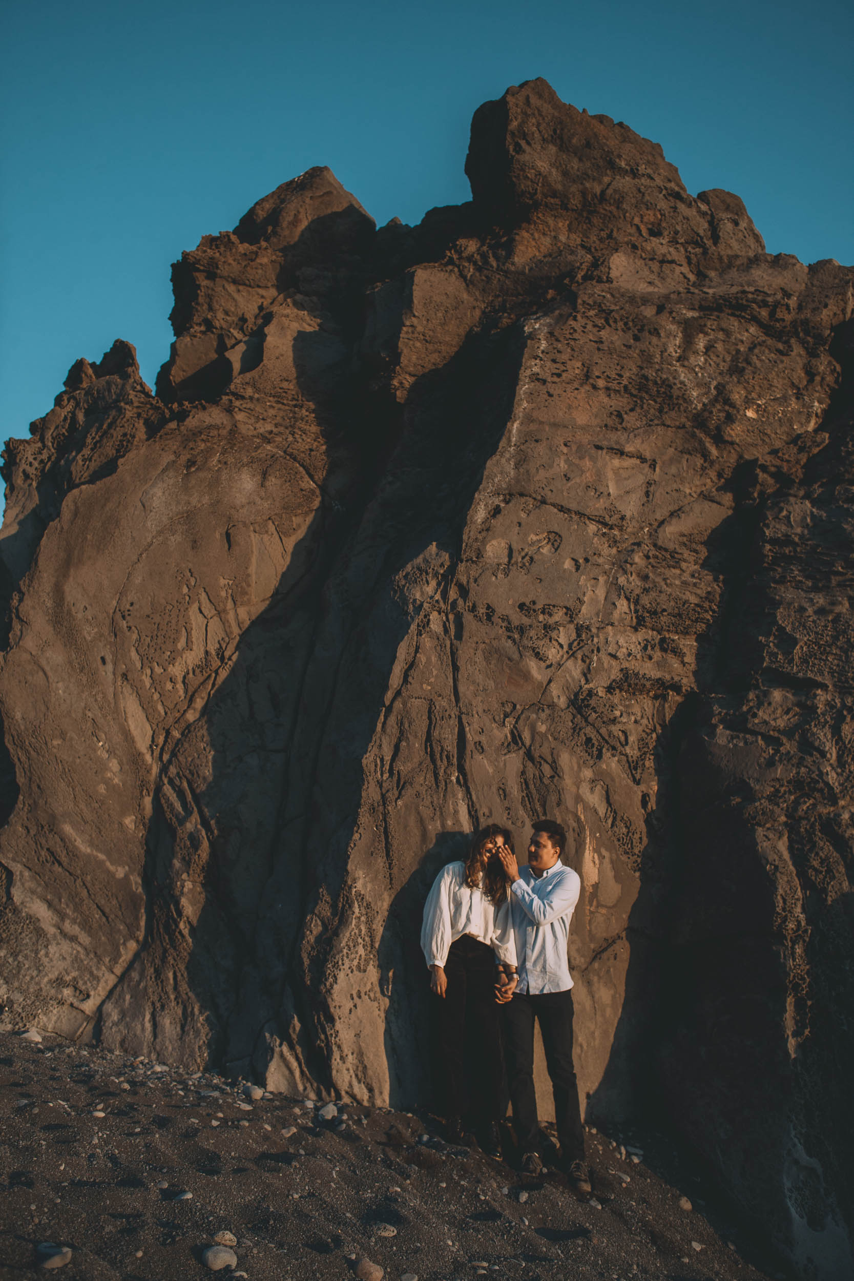 Couple standing in a narrow gap between tall volcanic rock formations at Praia Formosa, man gently holding her face, deep blue sky above, Madeira
