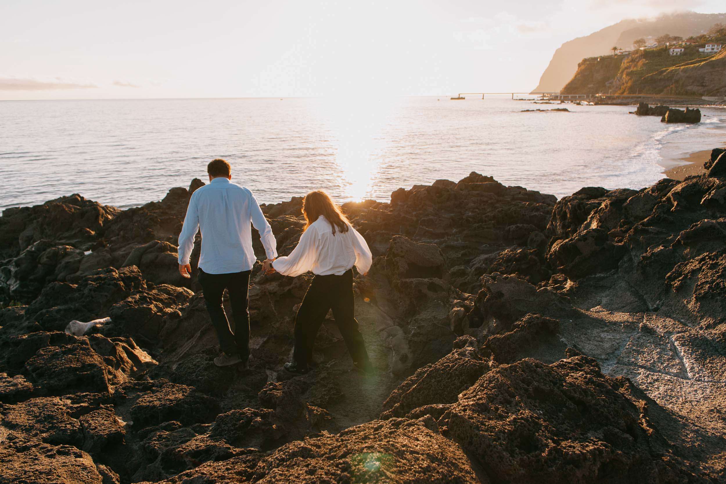 Couple walking hand in hand toward sunset, backs to camera, golden lens flare, Funchal cliffs and promenade behind, Praia Formosa, Madeira