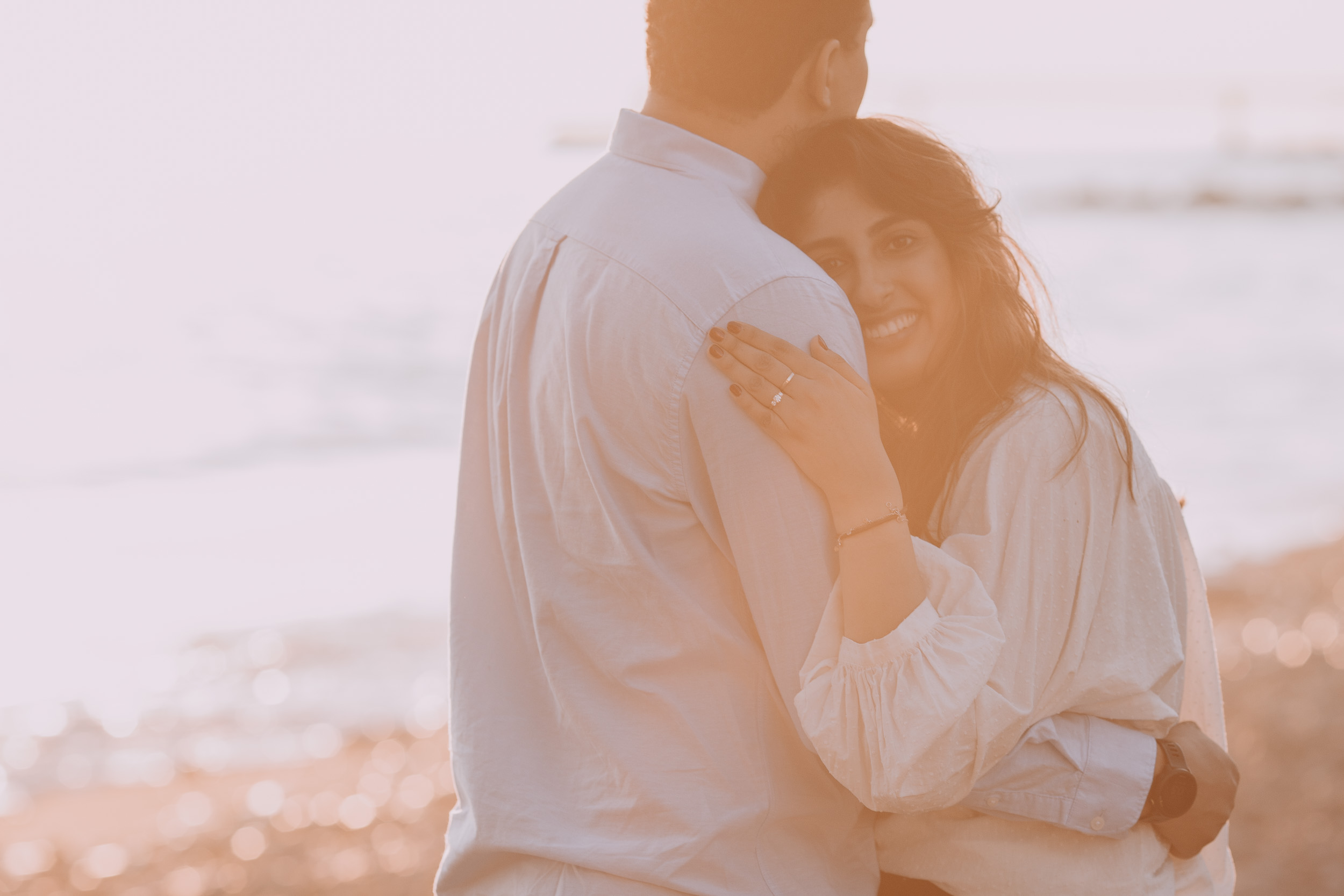 Woman hugging man from behind, engagement ring prominently visible against his shirt, she smiling, hazy golden backlight, Praia Formosa, Madeira