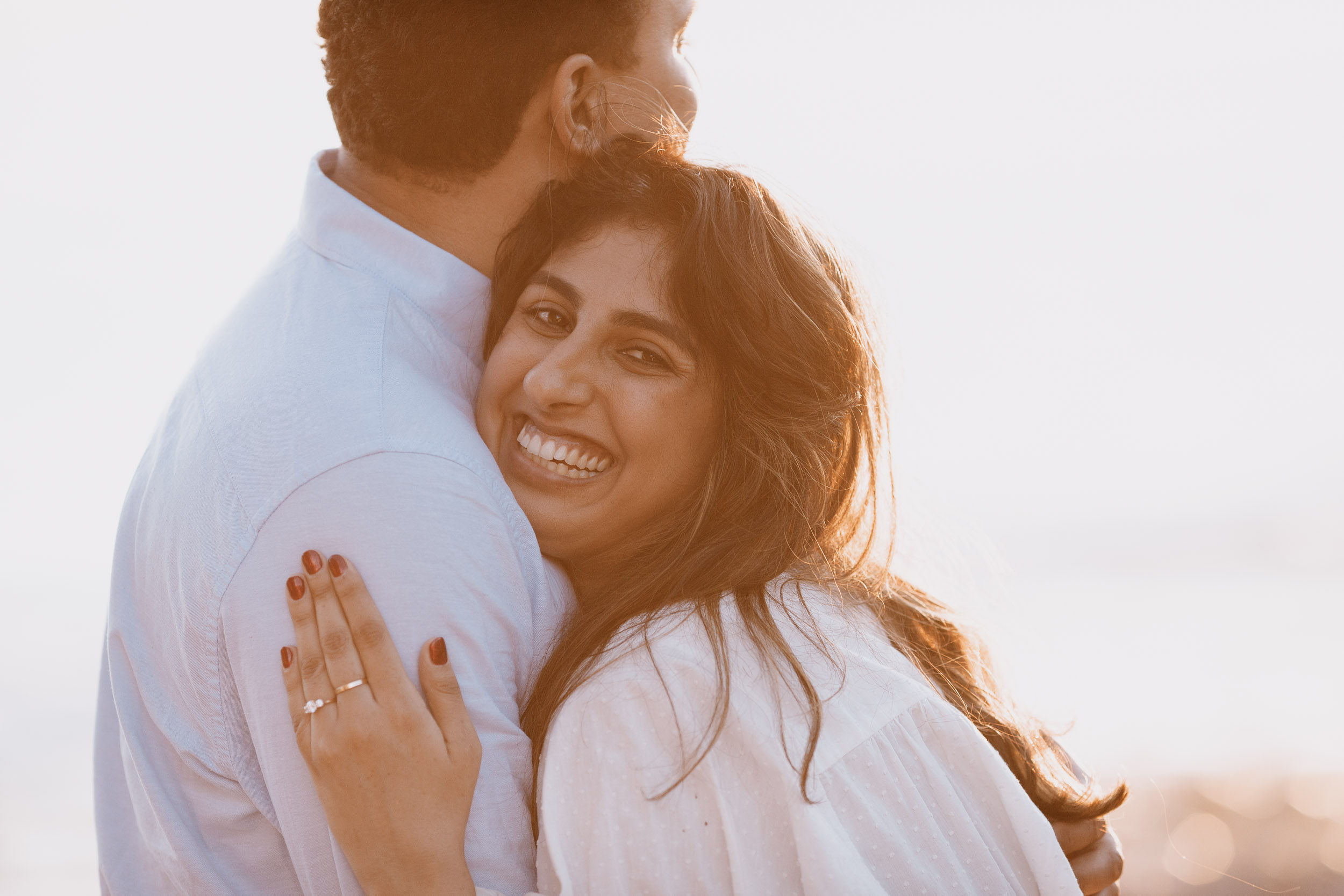Close-up of woman beaming pressed against man's shoulder, engagement ring clearly visible on hand, warm golden backlight, Praia Formosa, Madeira