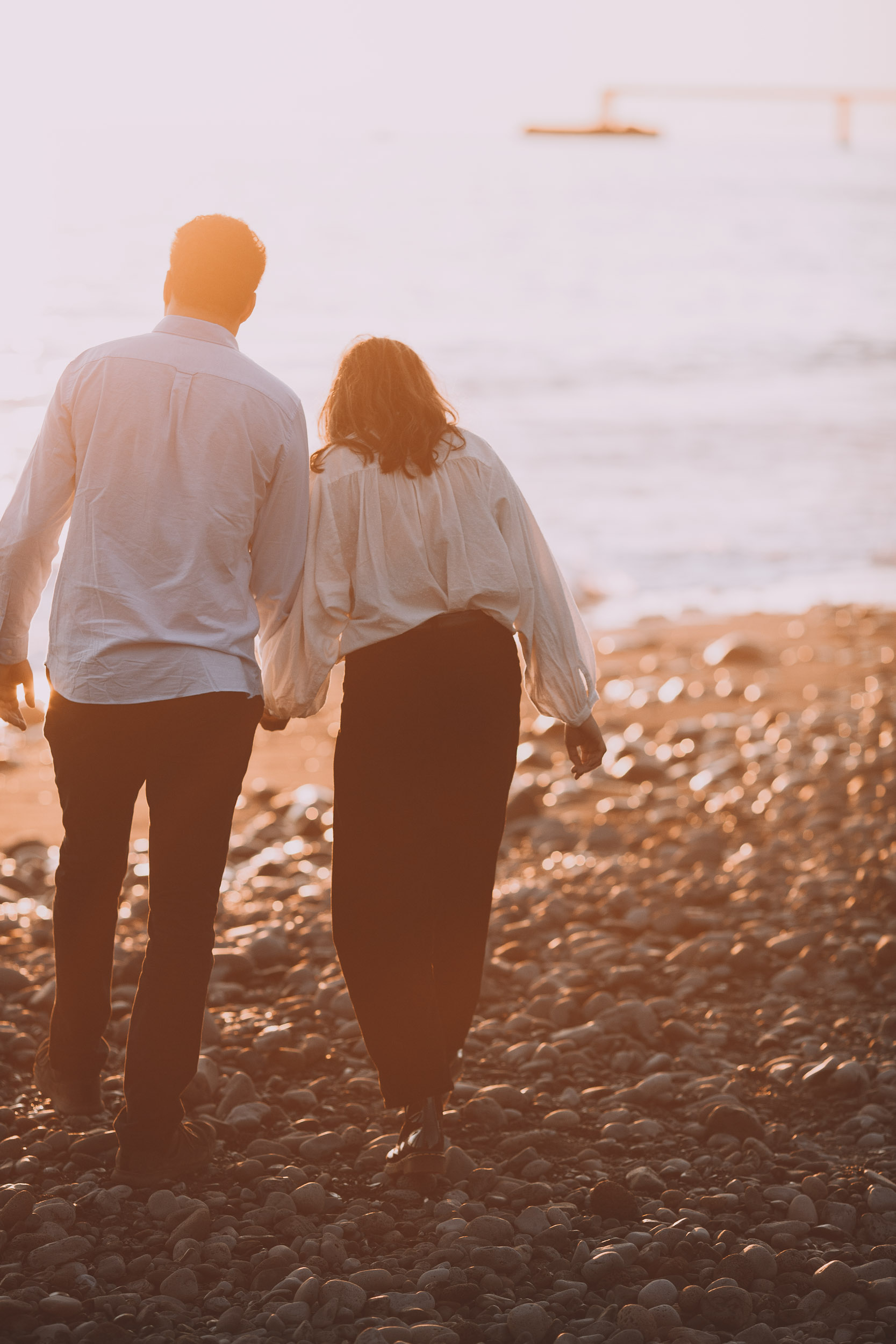 Couple walking hand in hand along pebble beach toward the Atlantic, backs to camera, golden sunset, Funchal promenade pier in distance, Praia Formosa, Madeira