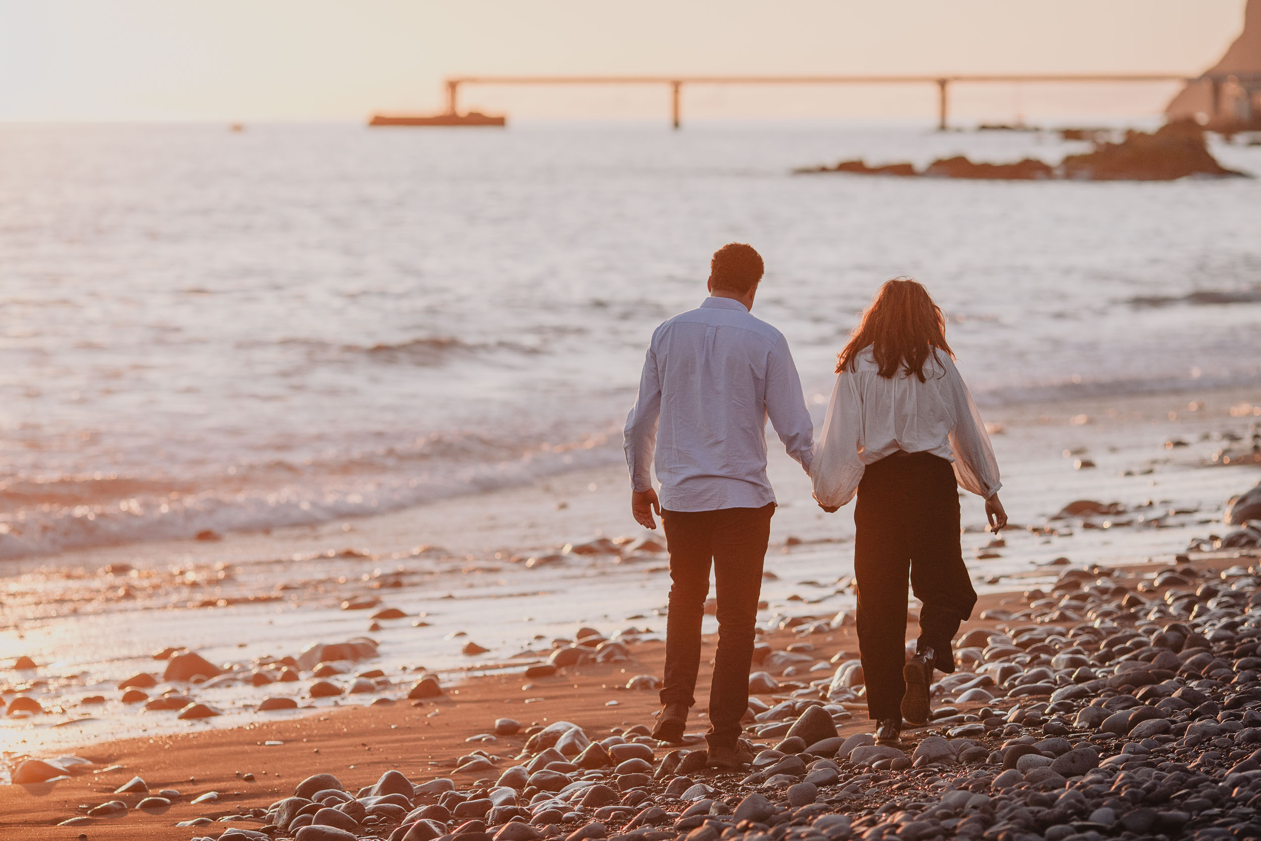 Couple walking along Praia Formosa pebble beach, backs to camera, Funchal bridge and promenade in distance, golden afternoon light, Madeira