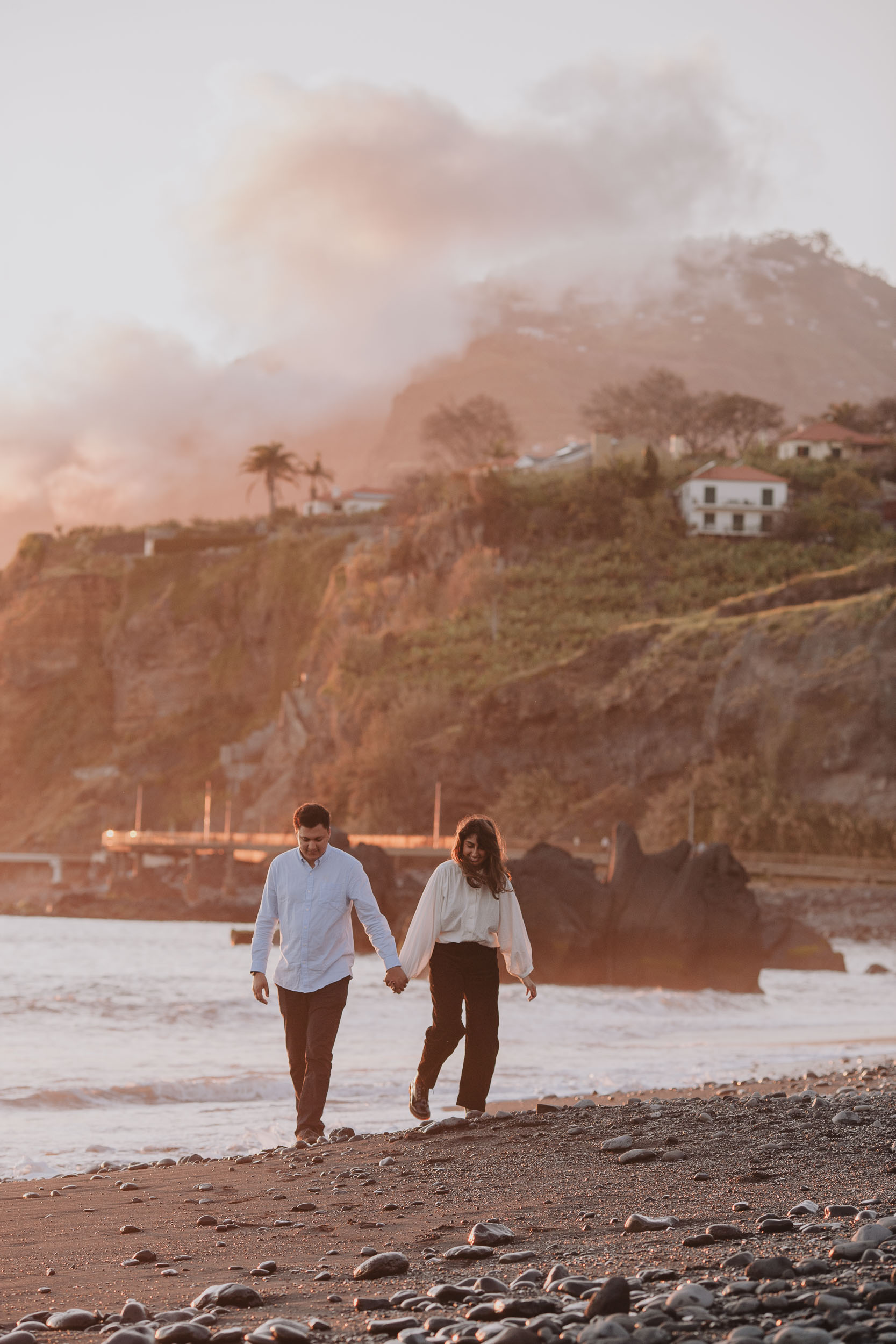 Couple kissing on Praia Formosa pebble beach, she cupping his face tenderly, Funchal cliffs with white houses behind in misty golden light, Madeira