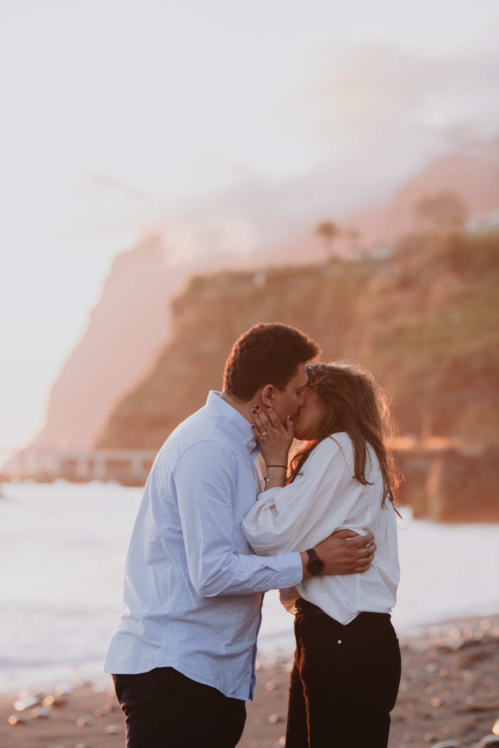 Woman looking over partner's shoulder toward camera, ring visible on her hand, pebble beach at Praia Formosa, Madeira
