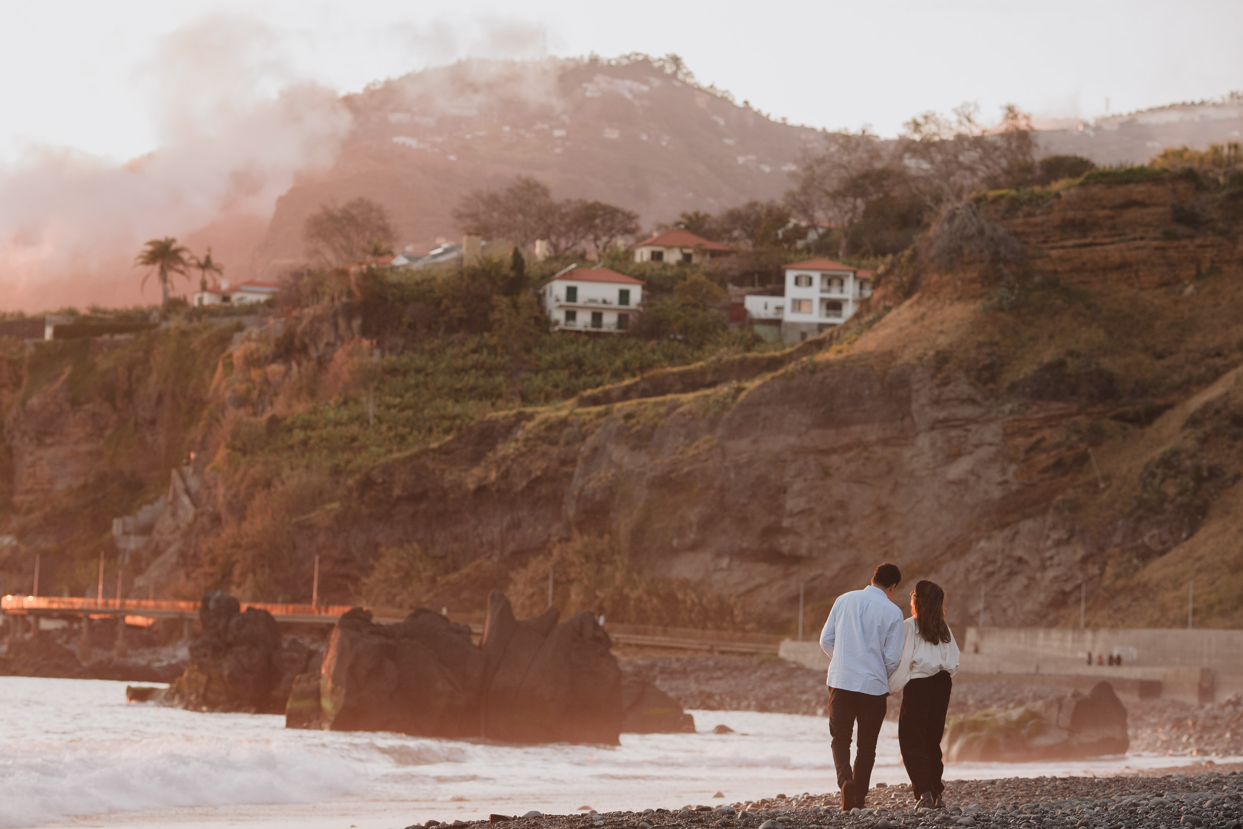 Couple walking together along Praia Formosa pebble beach, backs to camera, large Funchal hillside with houses and mist behind, Madeira