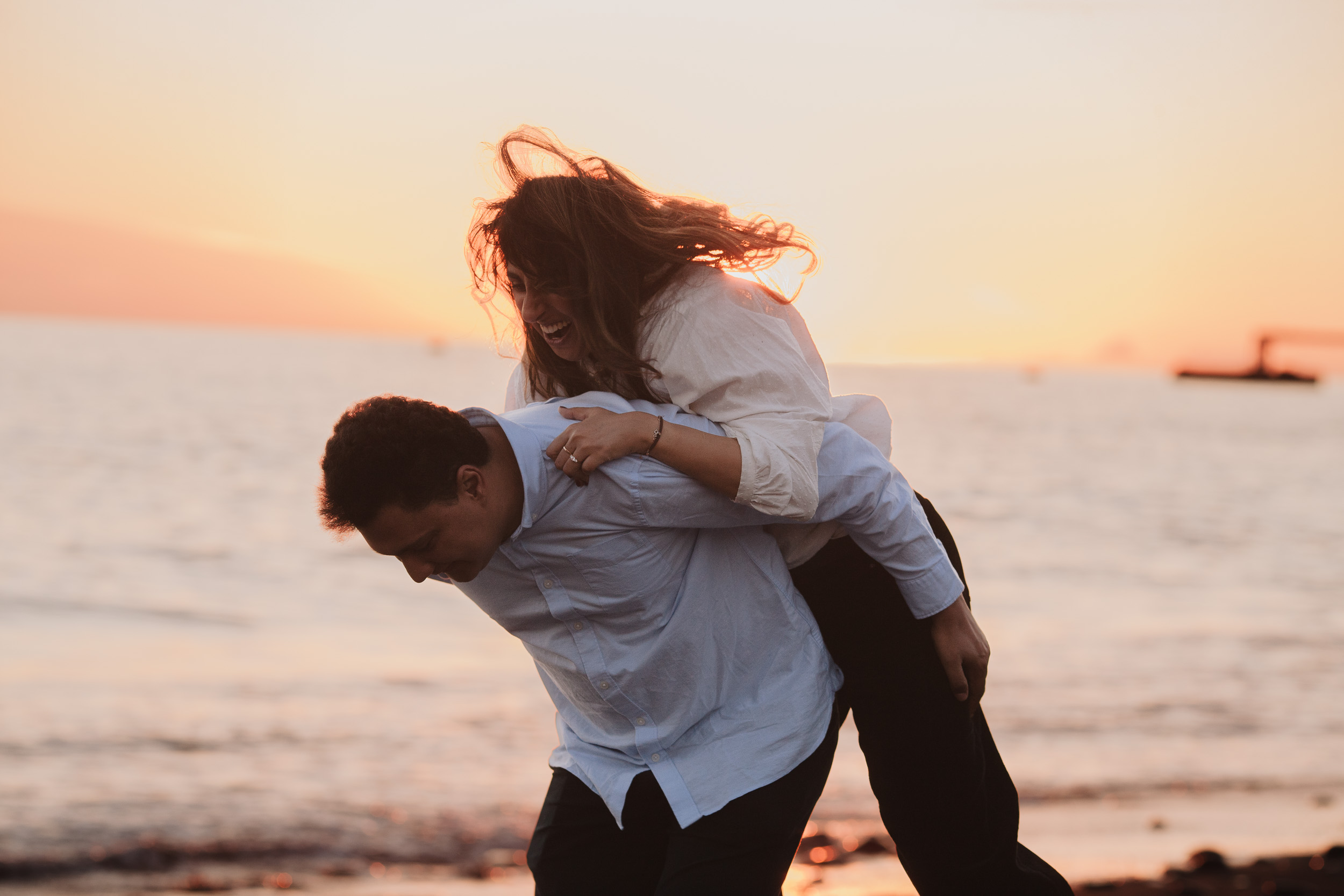Man carrying woman on his back at Praia Formosa, she laughing with hair flying in the ocean breeze, orange Atlantic sunset sky, Madeira