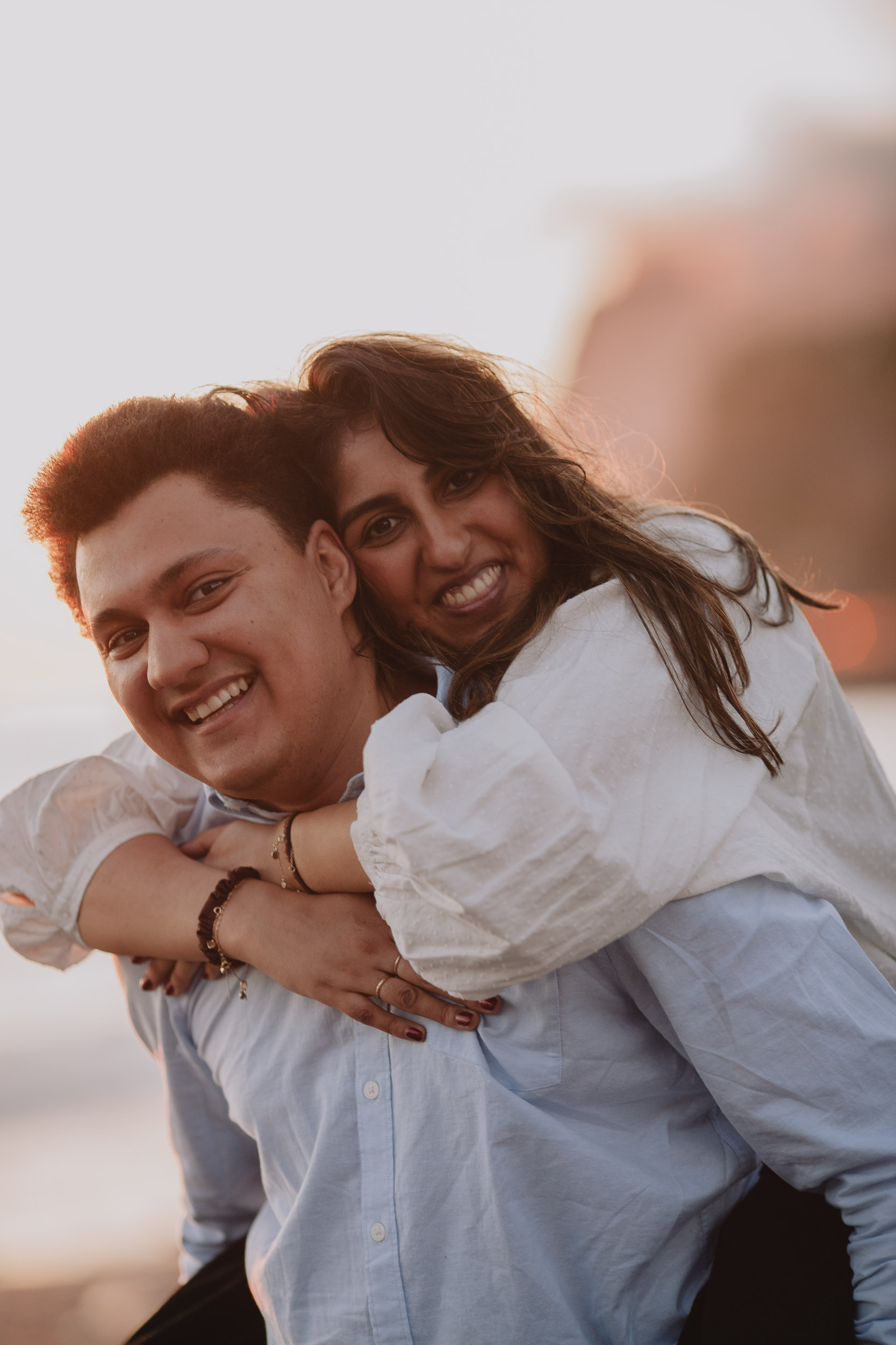 Both laughing close-up, she on his back with arms around his neck, engagement ring visible, warm golden backlight, Praia Formosa, Madeira