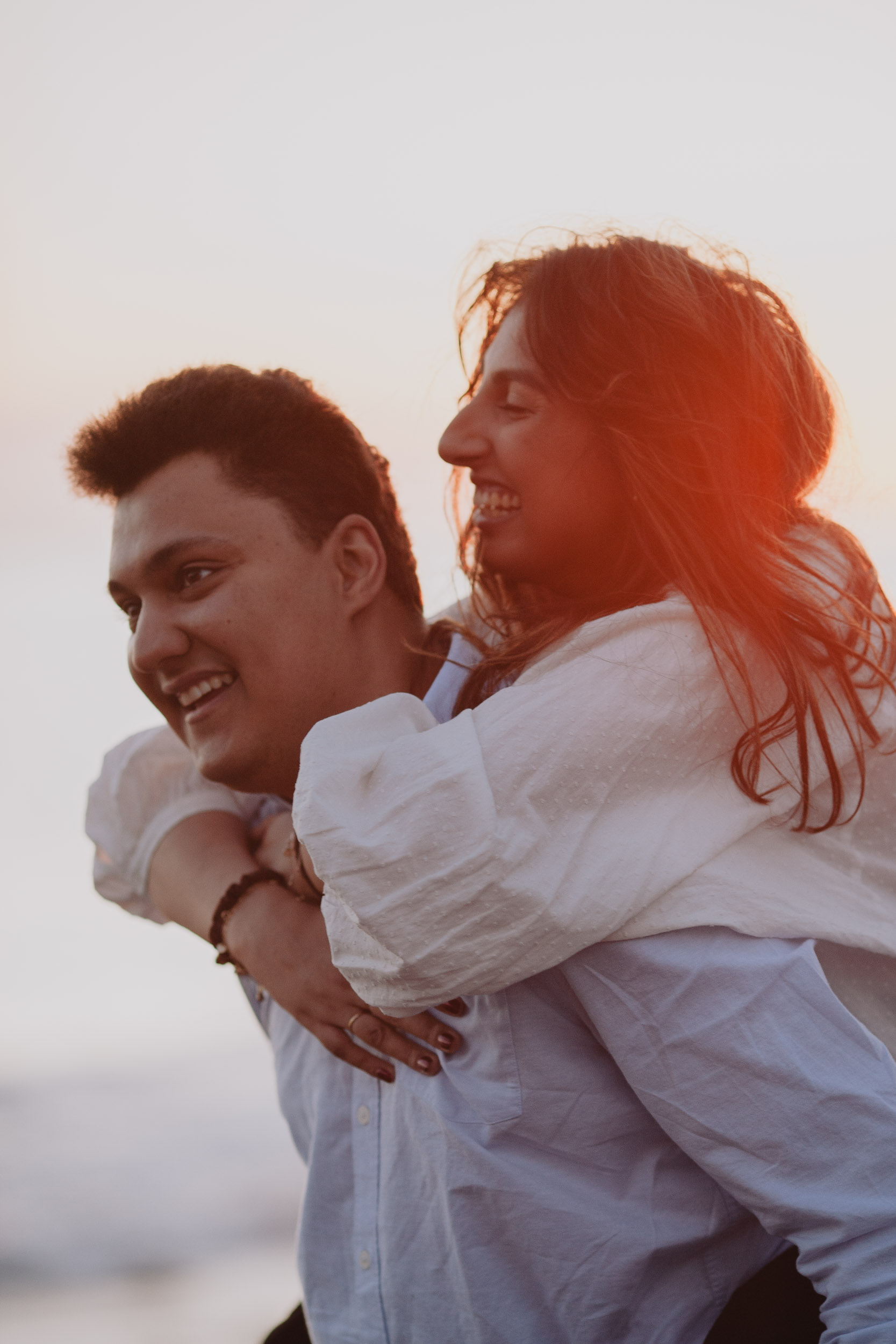 Couple laughing in profile, she on his back arms clasped around his neck, strong red-golden sunset flare behind, Atlantic horizon, Praia Formosa, Madeira