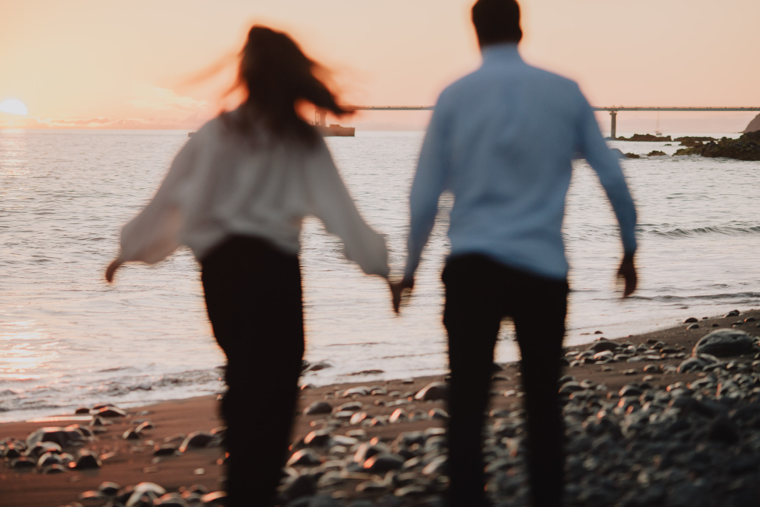 Couple running hand in hand along Praia Formosa pebble beach in motion blur, backs to camera, orange sunset sky, Funchal pier visible, Madeira