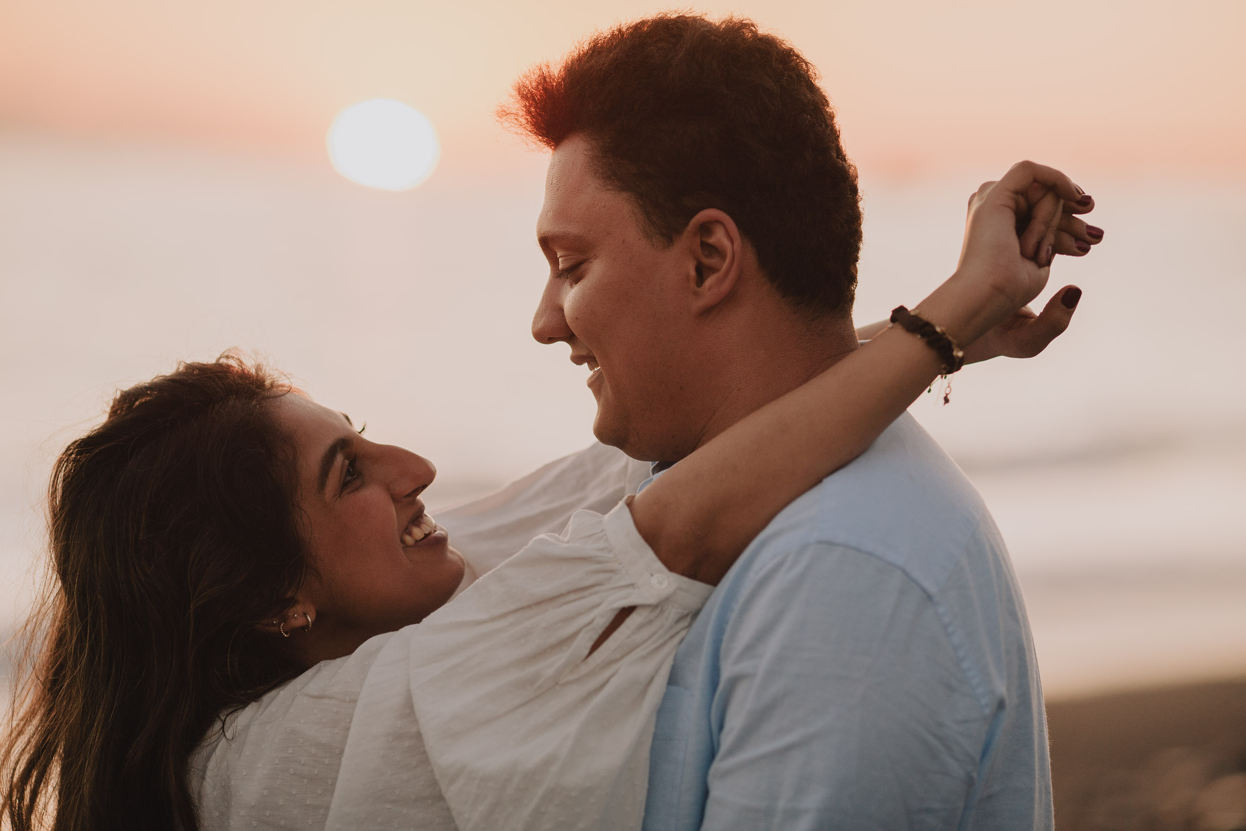 Couple smiling at each other closely, she with arm over his shoulder, full sun disc on Atlantic horizon, pink sunset sky, Praia Formosa, Madeira